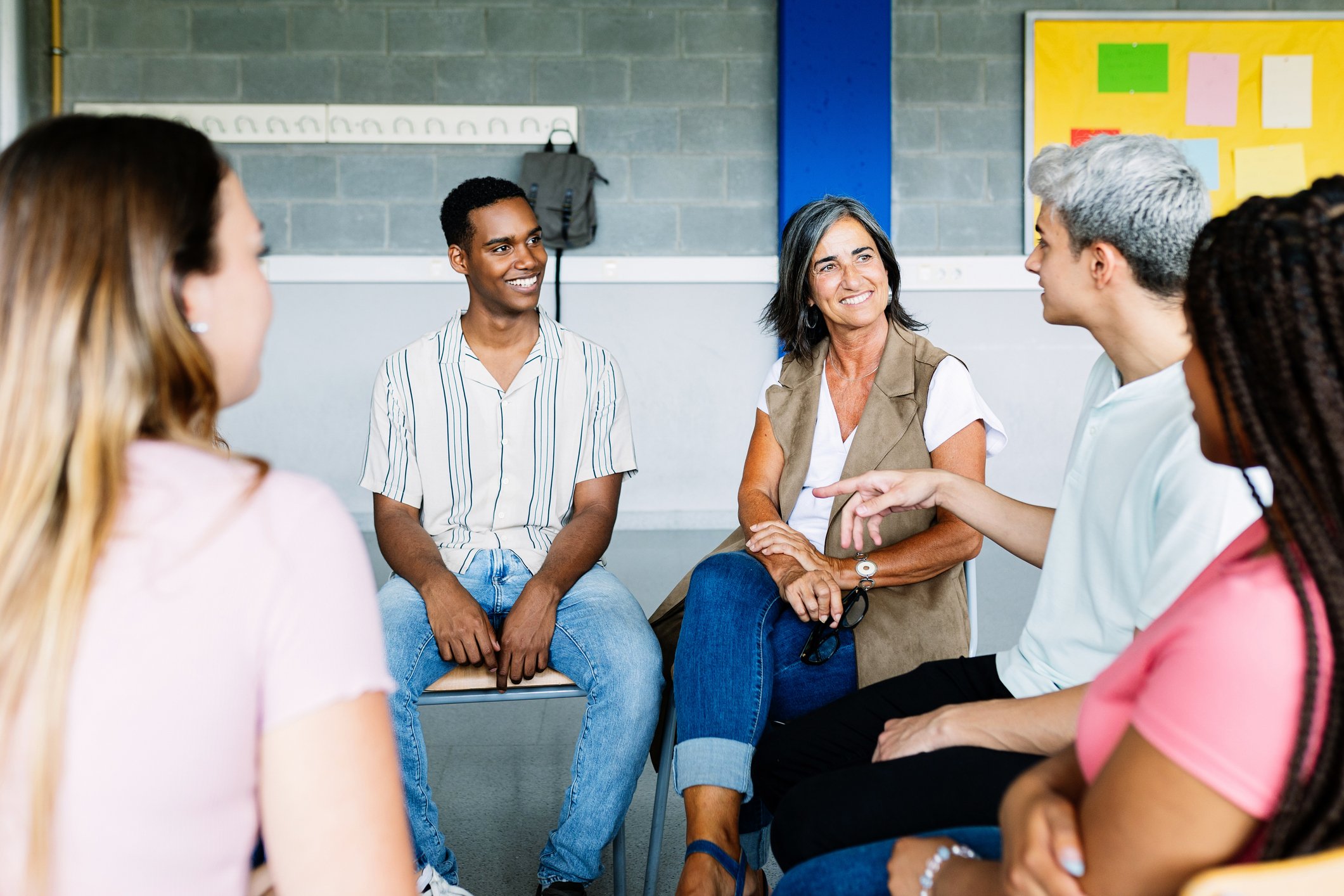 Group of diverse young women sitting in a semi-circle, engaging in a discussion with an older woman in a classroom or meeting room.