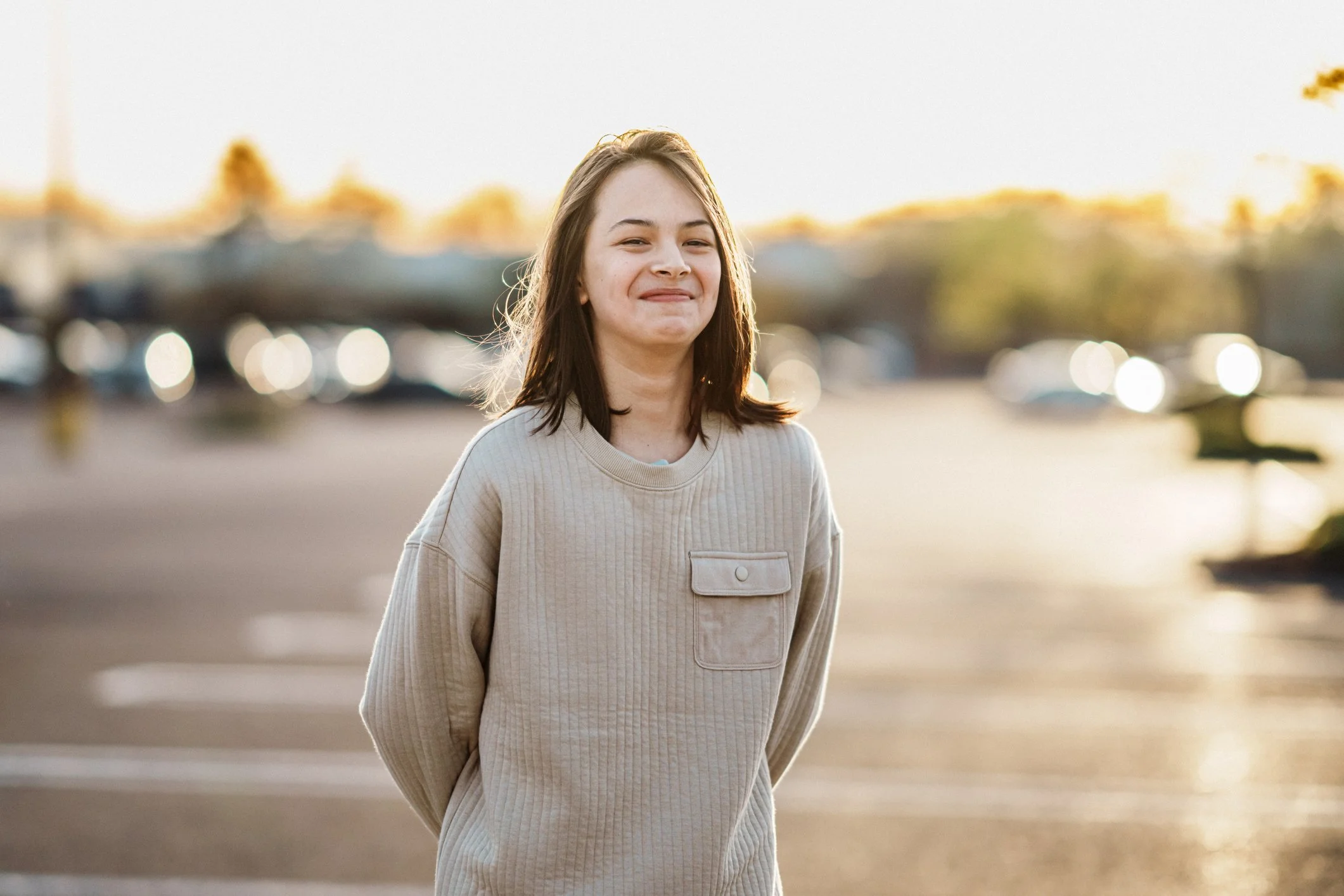 A young woman with light brown hair in a beige sweatshirt standing outdoors during sunset.