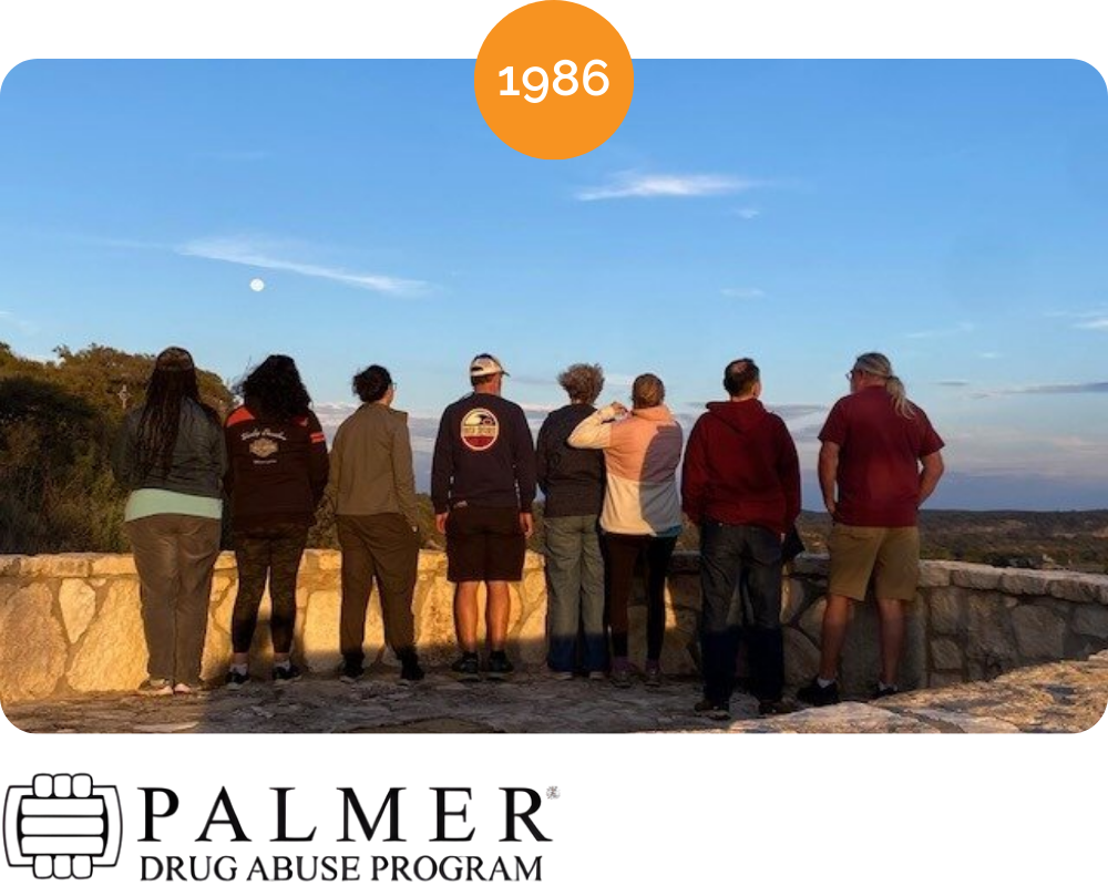 A group of nine people standing by a stone railing, watching the sunset or sunrise outdoors with trees and open sky in the background, and a full moon visible in the sky.
