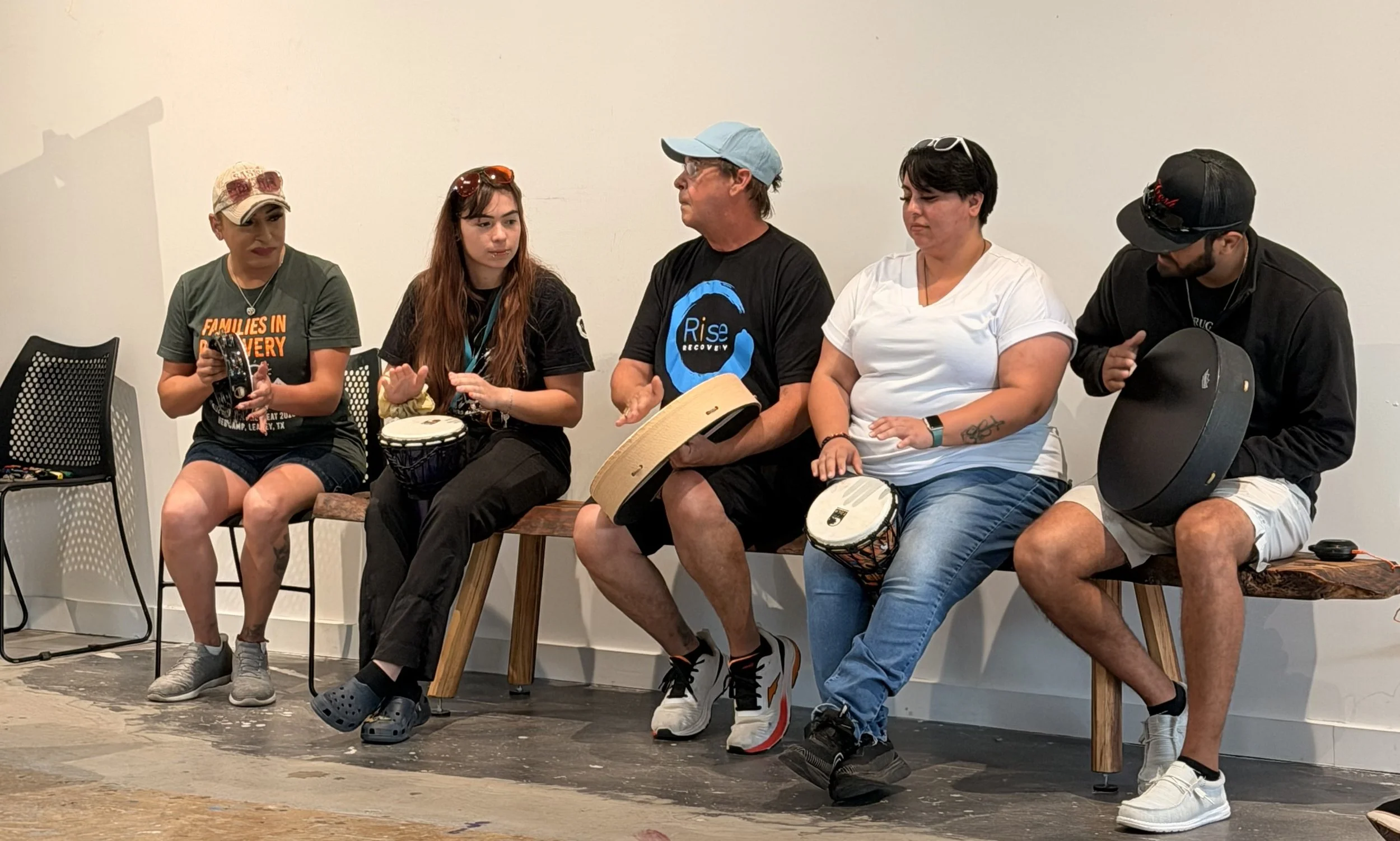 Five people sitting on chairs and a stool, engaging in drumming in a room with a plain wall background.
