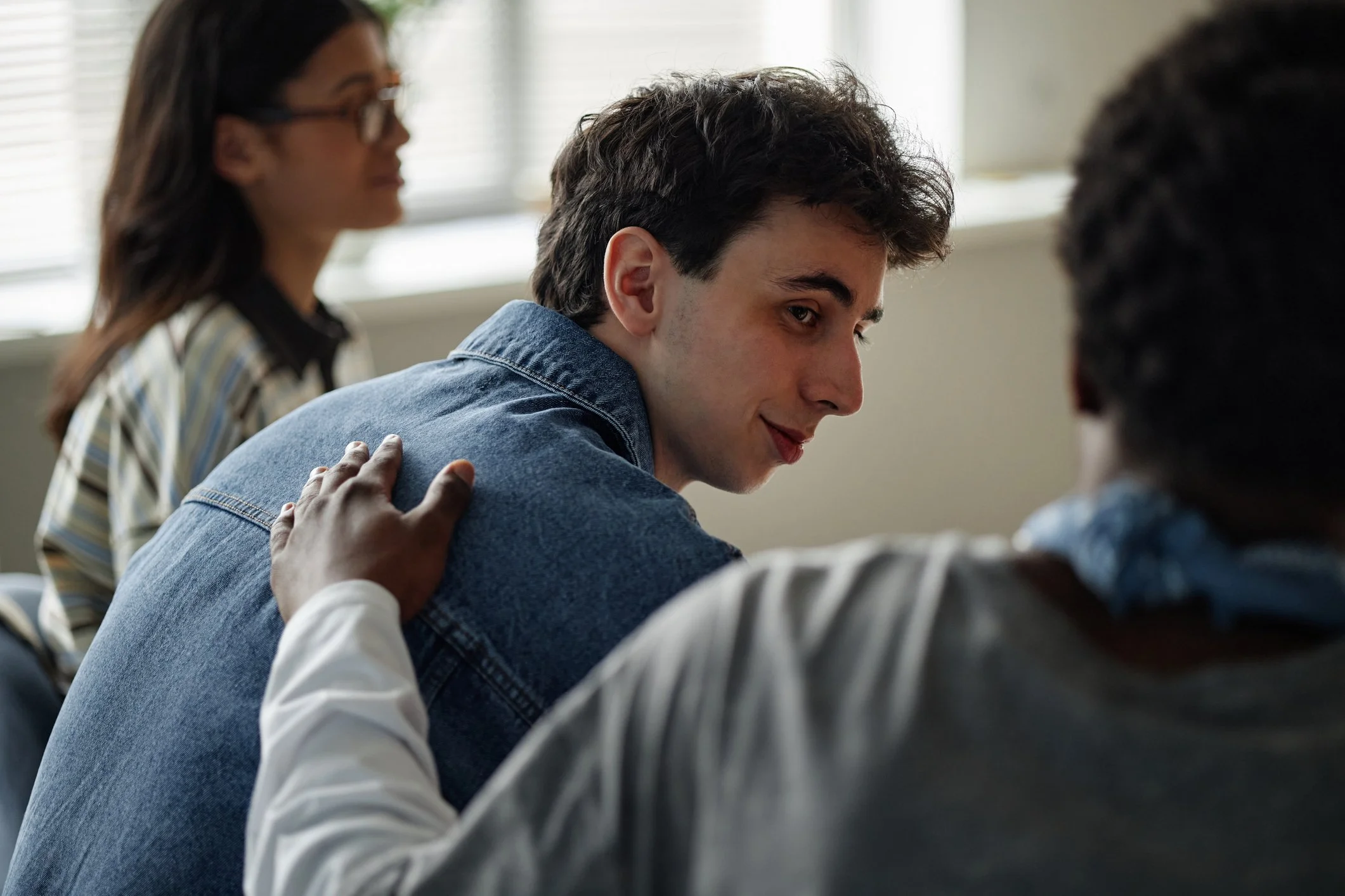 People gathered in a classroom, with focus on a young man with dark, curly hair smiling and looking to his right, while other individuals are partially visible and engaged in conversation.