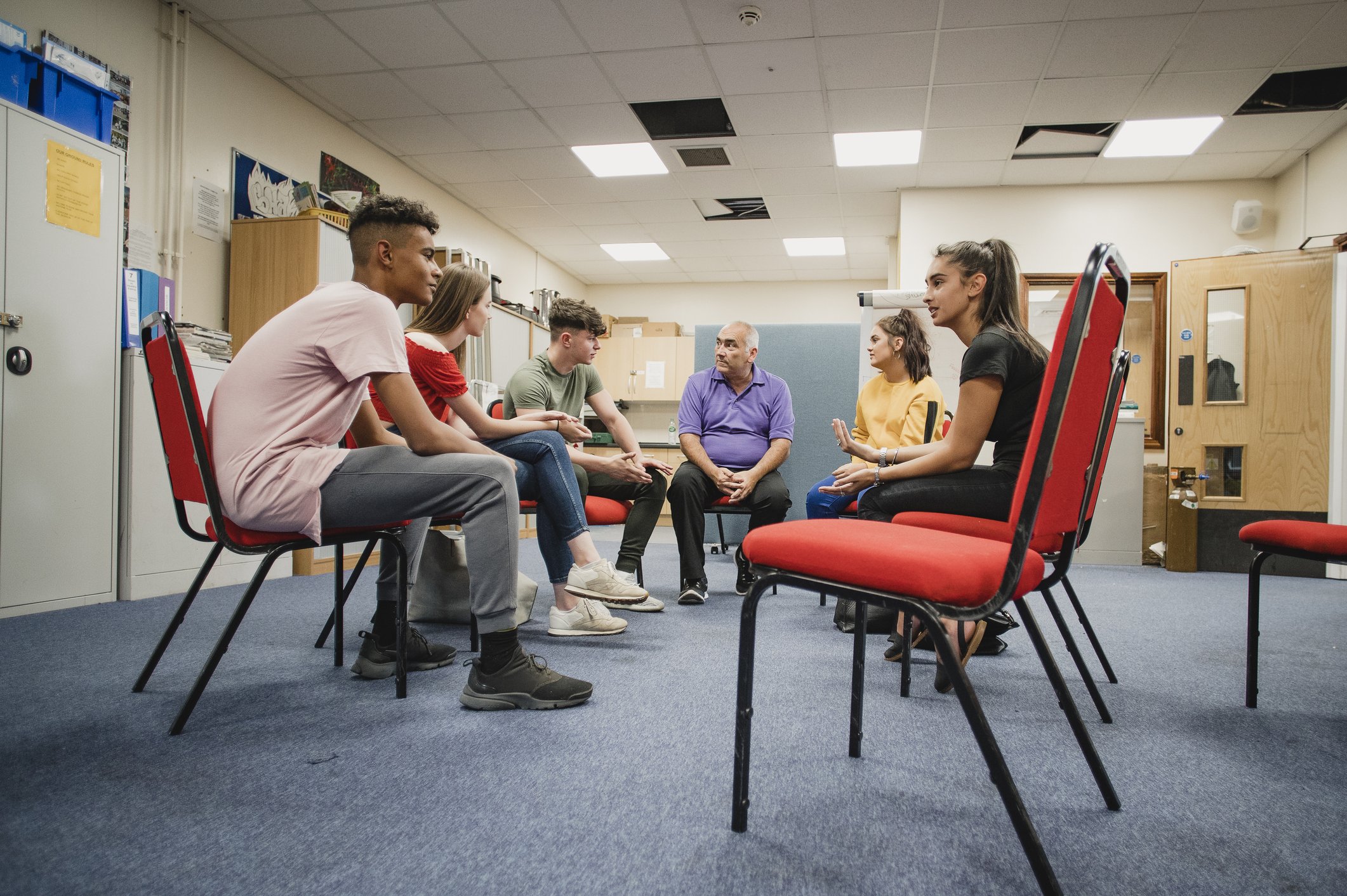 Six young adults sitting in a circle, engaged in a group discussion in an office or classroom setting with ceiling tiles, fluorescent lighting, and storage cabinets in the background.