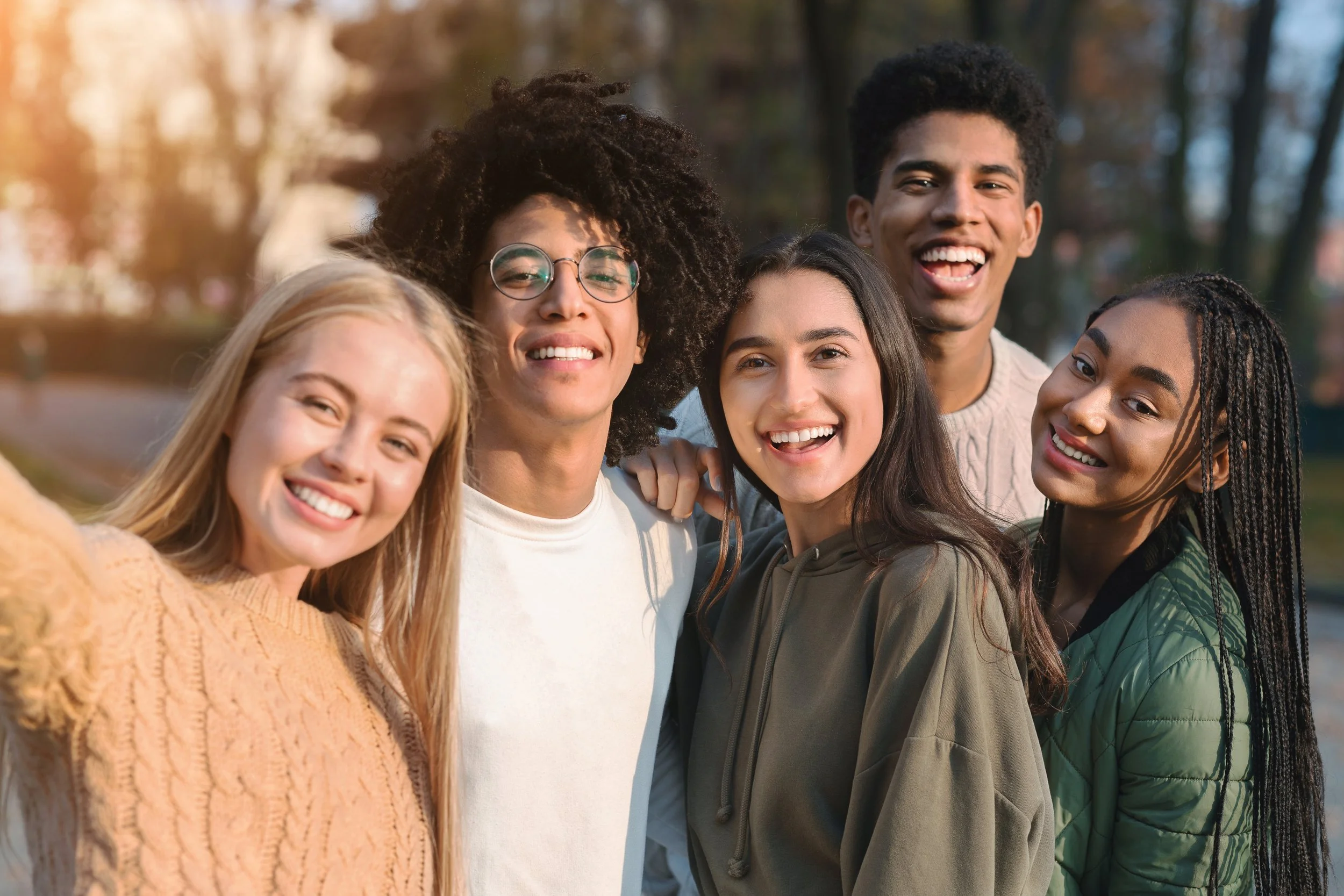 Group of six diverse young people smiling and taking a selfie outdoors in a park during autumn.