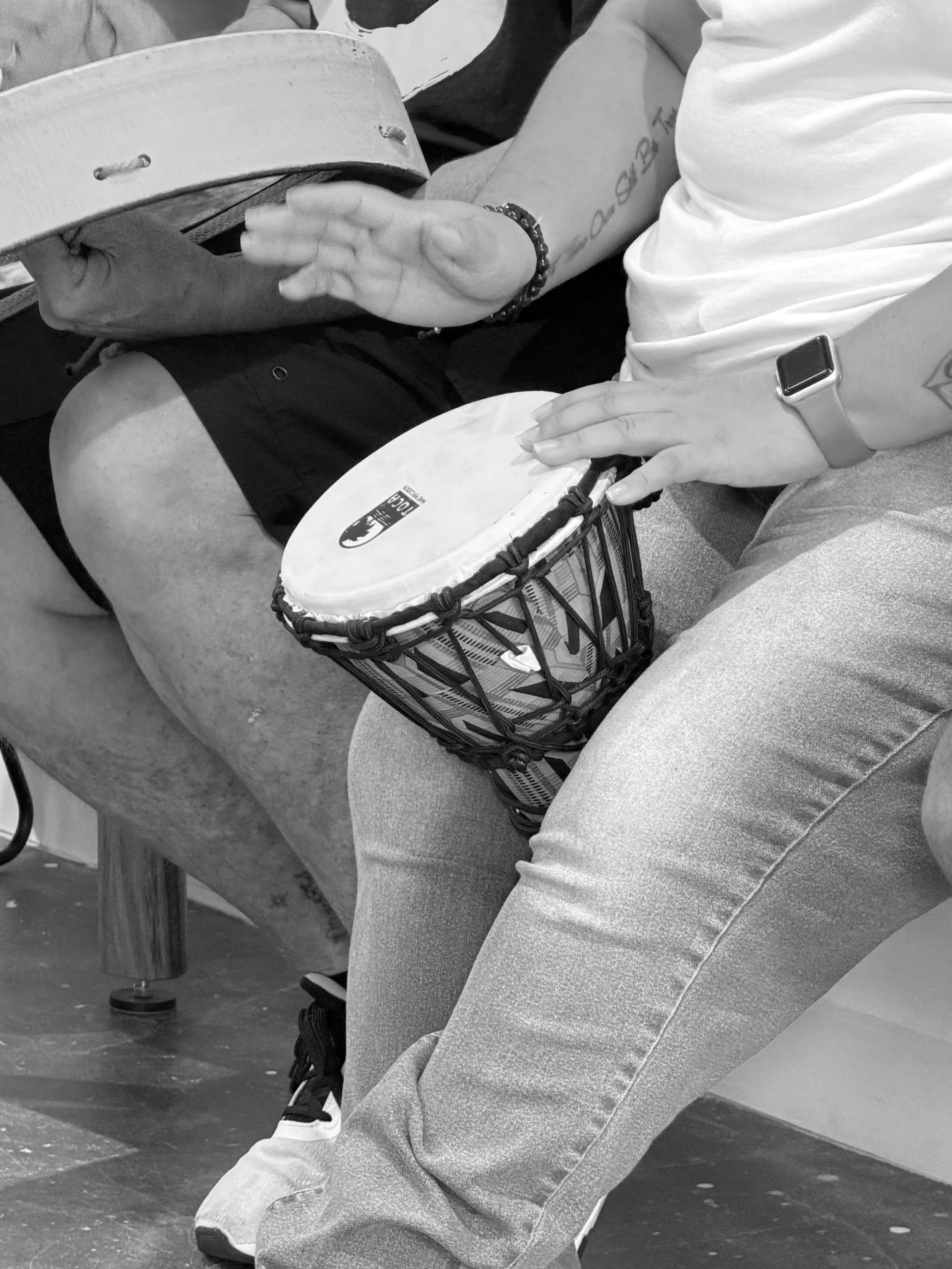 A person sitting with their legs crossed, holding a drum in their lap, wearing a white T-shirt, gray pants, and a wristwatch, with visible tattoos on their arms. Their other hand is resting on the drum.