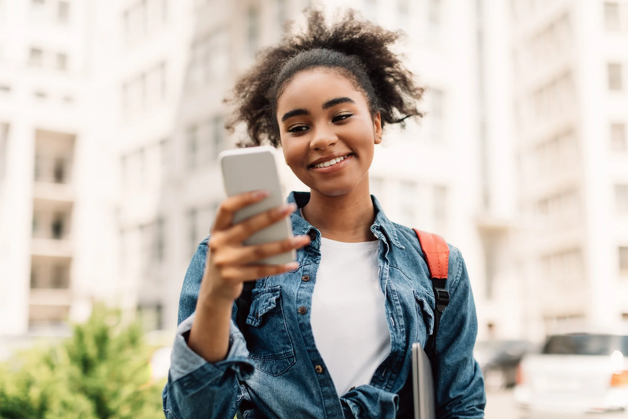 Young woman with curly hair and a denim jacket looking at her phone, smiling outdoors with apartment buildings in the background.