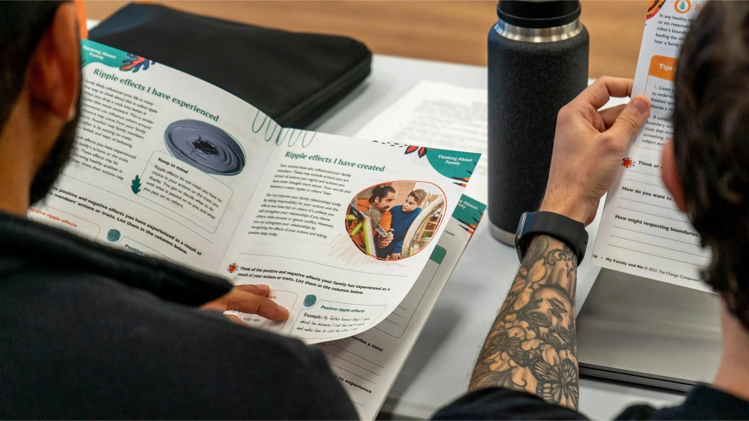 Two people sitting at a table, looking at a colorful informational brochure about ripple effects and family relationships. One person's arm with a floral tattoo is visible, and the other person has glasses. There is a black water bottle on the table, along with other papers and a black folder.