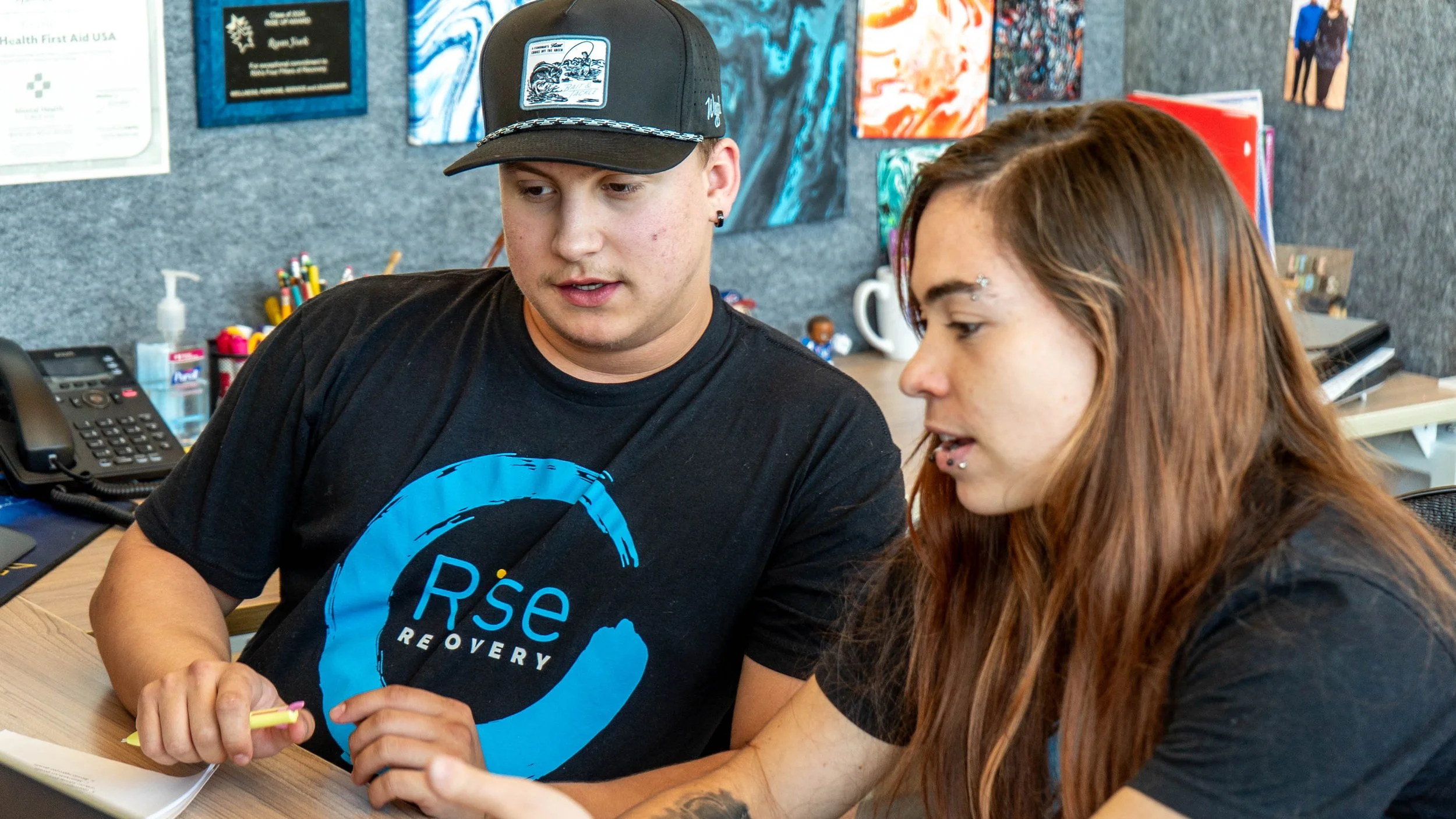 Two young adults, a man and a woman, sitting at a desk in an office, looking at something together. The man wears a black baseball cap with a patch and a black T-shirt with a blue circular logo that says 'Re' and 'Recovery'. The woman has long reddish-brown hair, facial piercings, and is wearing a dark top. The desk has a telephone, a hand sanitizer, and various office supplies. The background features gray walls with colorful abstract art and posters.