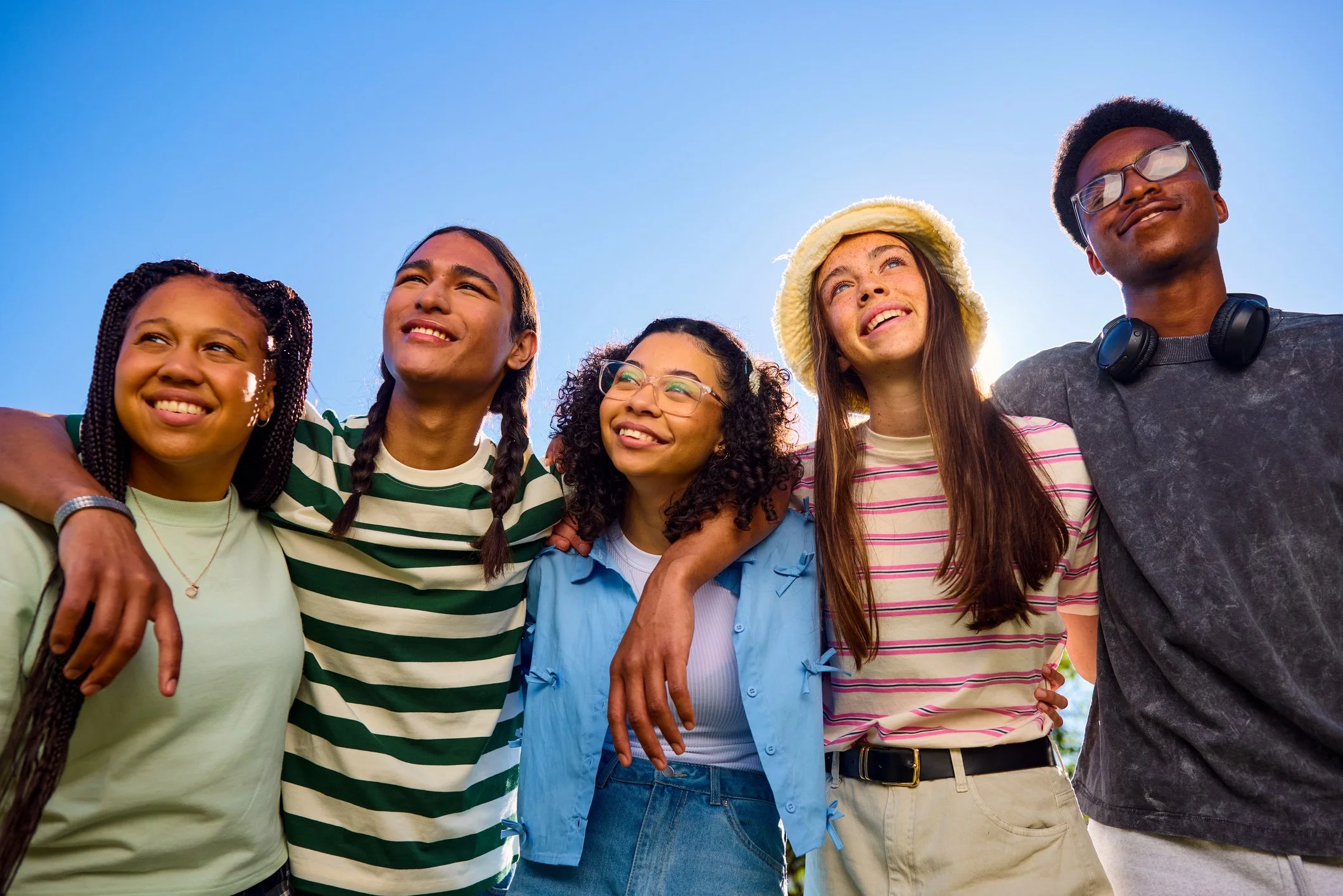 Group of five young friends standing close together outside under a clear blue sky, smiling and looking up, with their arms around each other.