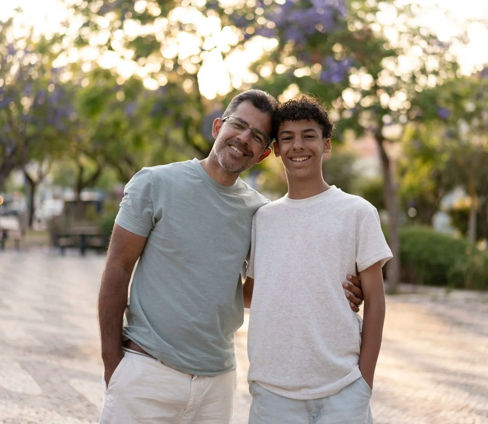 A man and a boy smiling and standing close together outdoors with trees in the background during sunset.