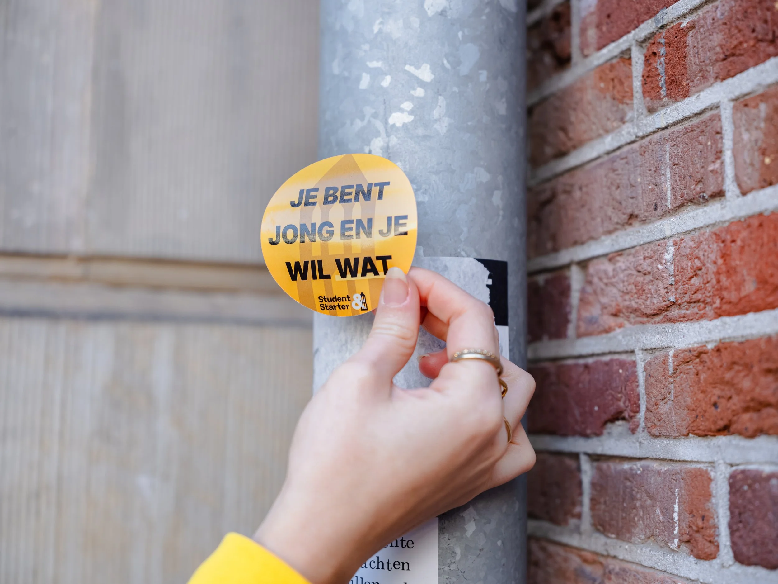 Een hand plakt een sticker op een regenpijp met de tekst "Je bent jong en je wil wat"