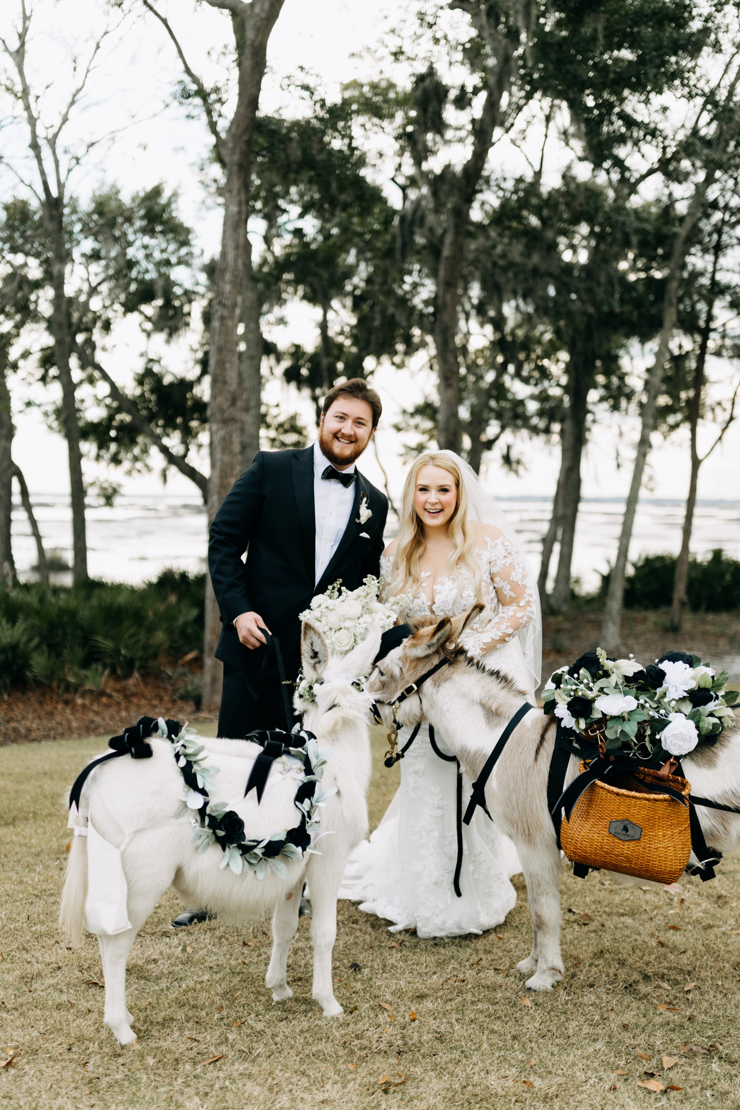 A joyful couple in wedding attire standing outdoors near the lake, holding two decorated donkeys with floral arrangements.