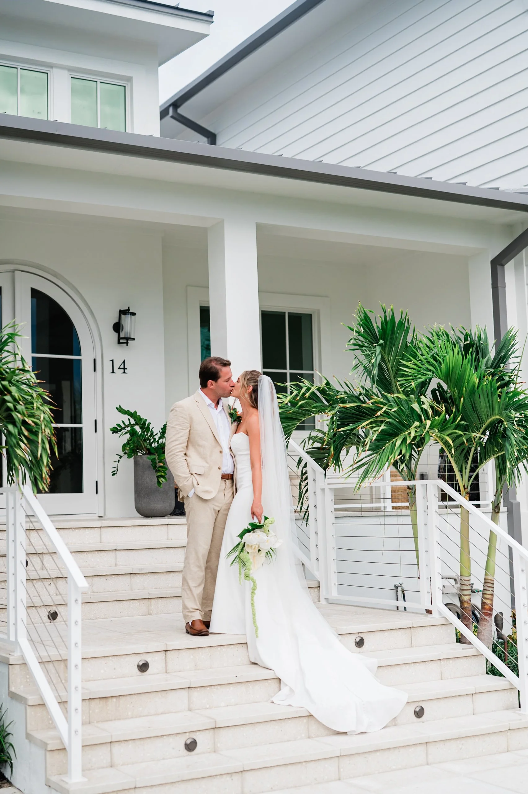 A bride and groom kissing on the steps of a white modern house, surrounded by tropical plants.