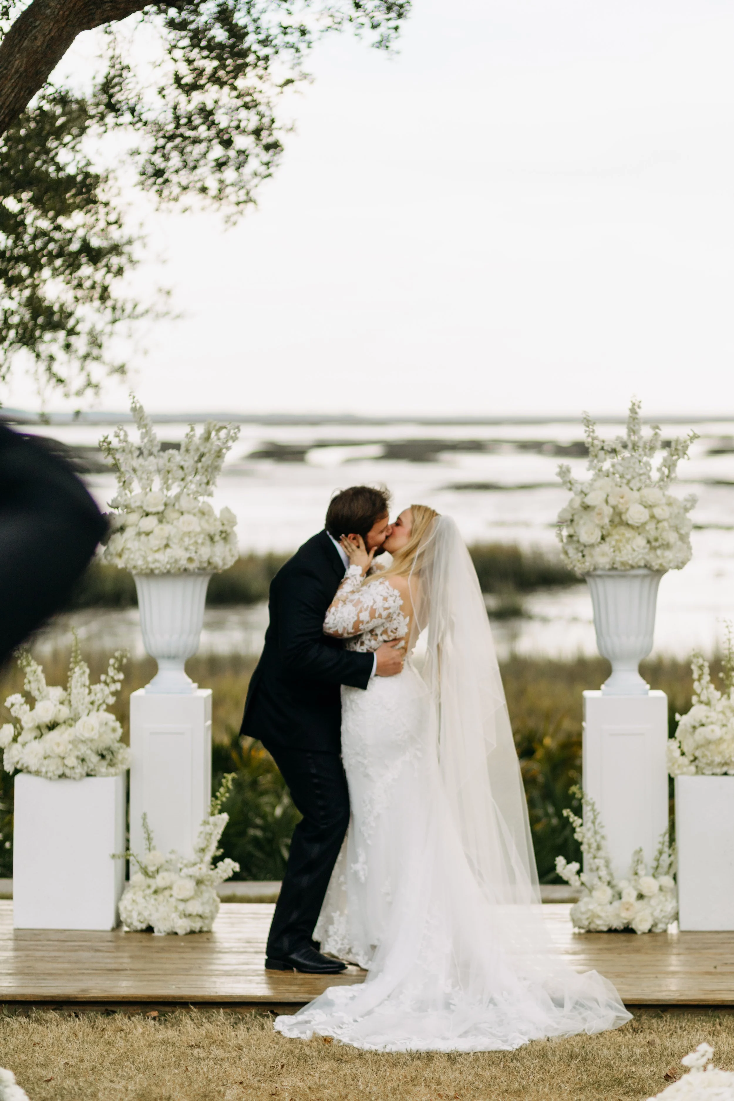 A bride and groom sharing a kiss during their outdoor wedding ceremony, with white floral arrangements on tall vases and a scenic waterfront background.