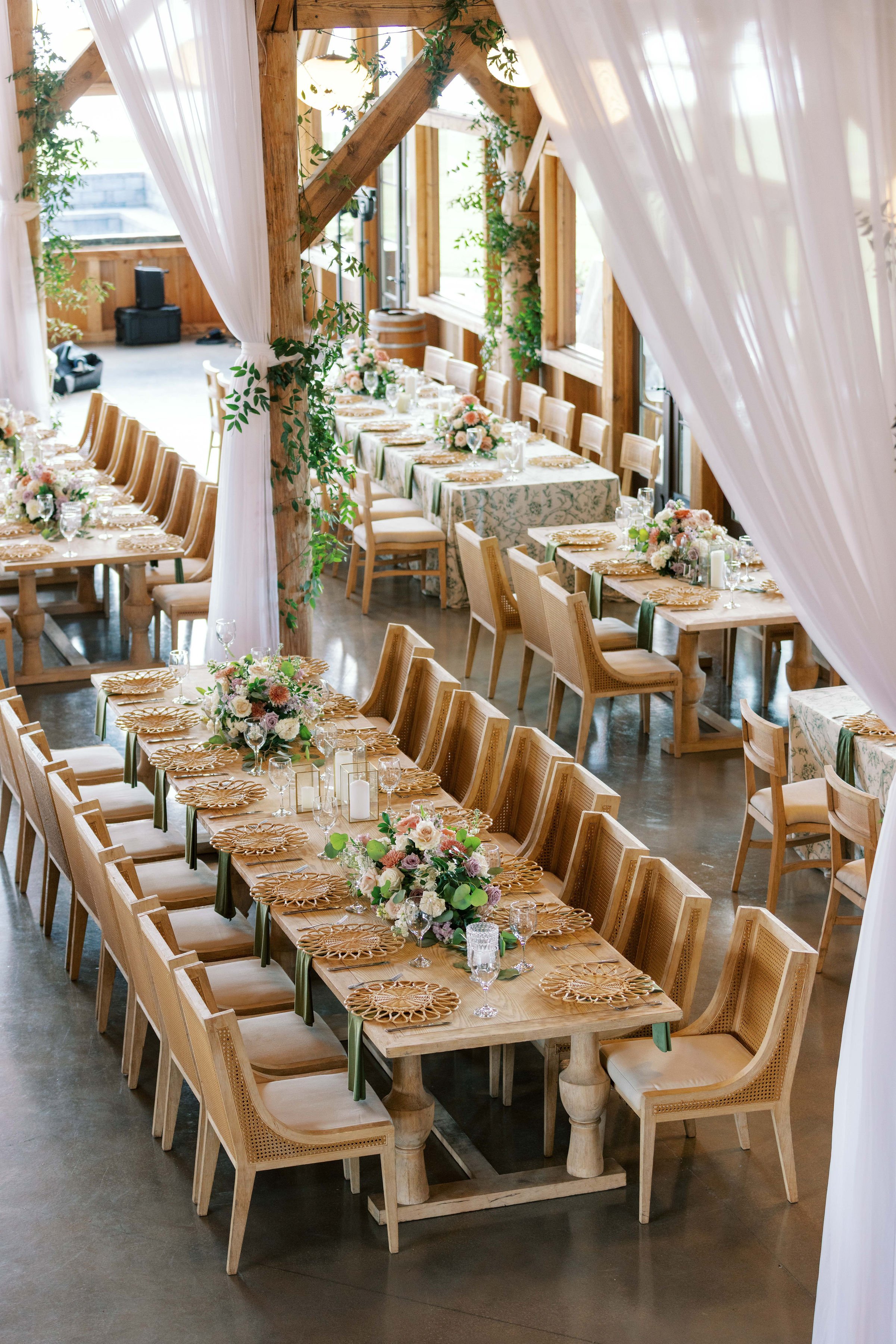 Elegant wedding or event dining table setup with floral centerpieces, candles, glassware, and gold chargers in a rustic wooden venue with high ceilings and flowing white curtains.
