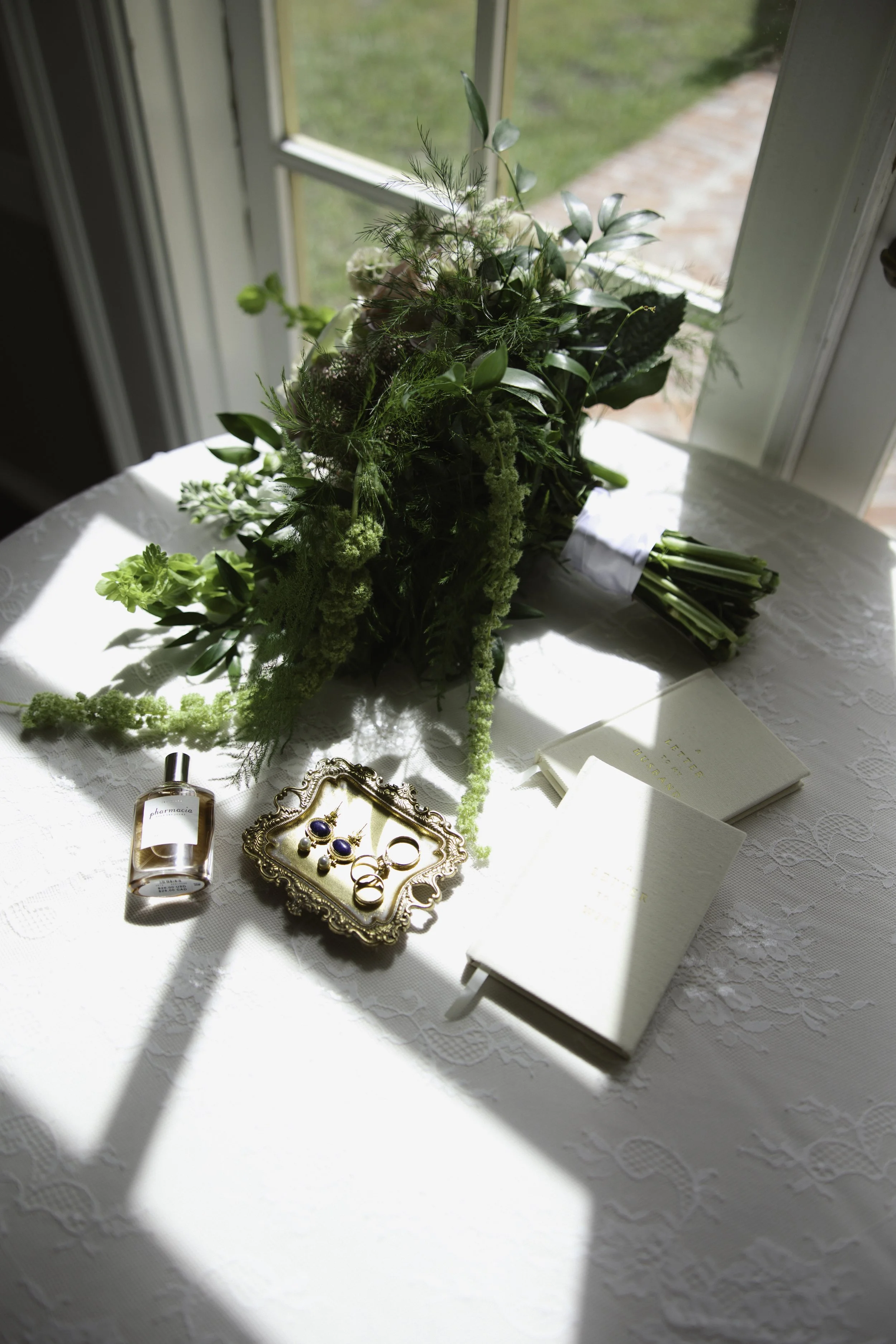 A bouquet of green and white flowers on a white lace tablecloth, with jewelry, perfume, and greeting cards nearby, near a sunny window.