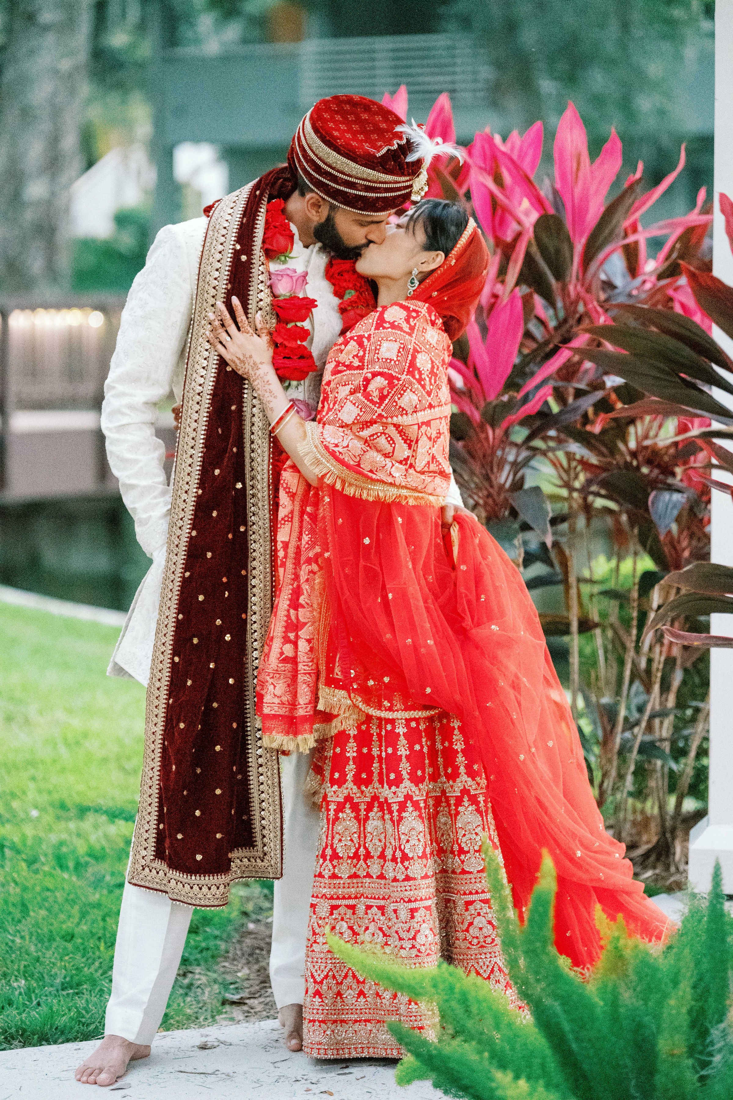 A couple dressed in traditional Indian wedding attire sharing a kiss outdoors, with pink and green plants in the background.