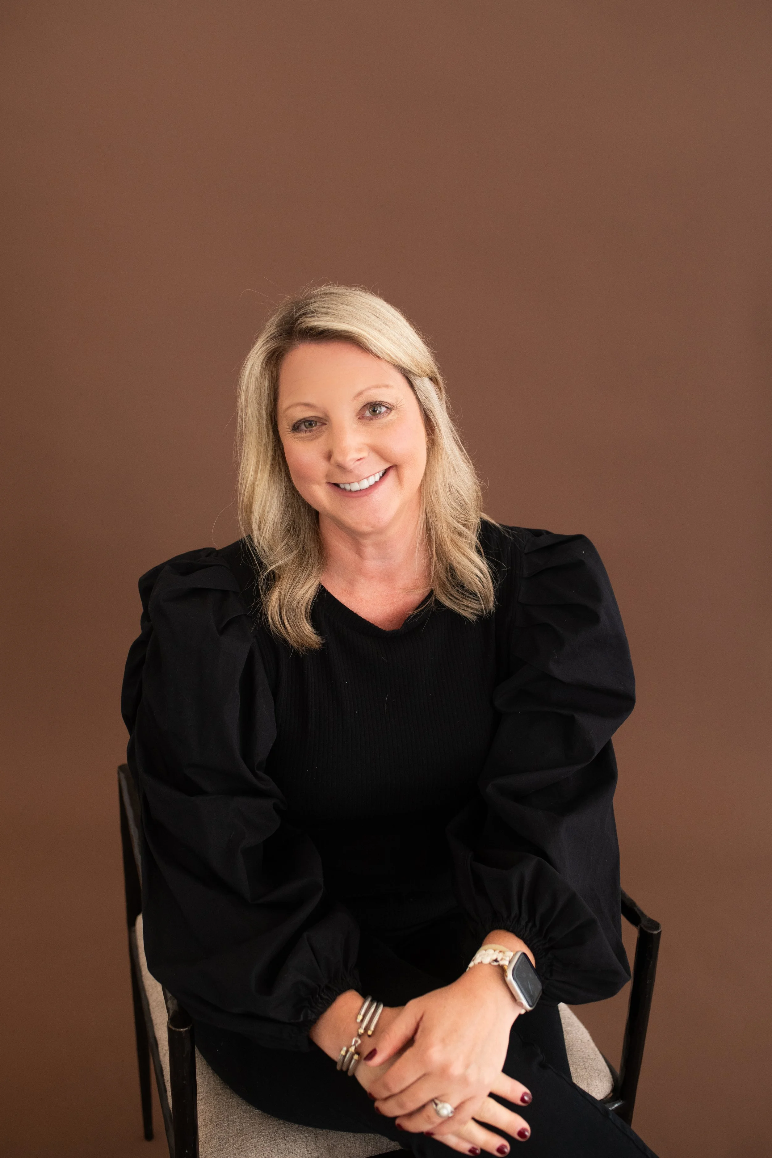 A woman with blonde hair wearing a black top with puffed sleeves, sitting on a chair against a brown background, smiling and looking at the camera.