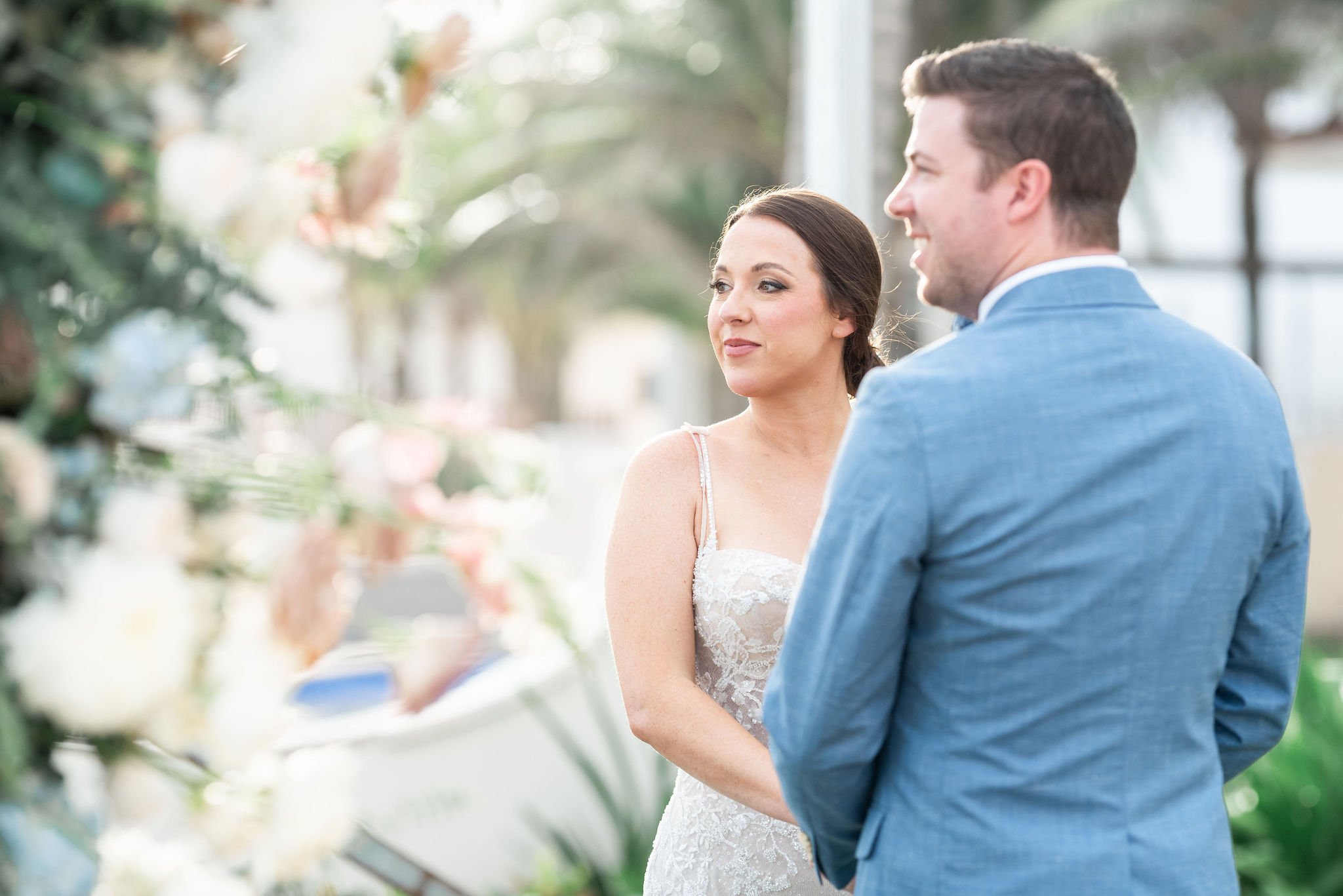 A bride and groom standing face to face outdoors during their wedding ceremony, surrounded by soft natural light and blurred greenery in the background.
