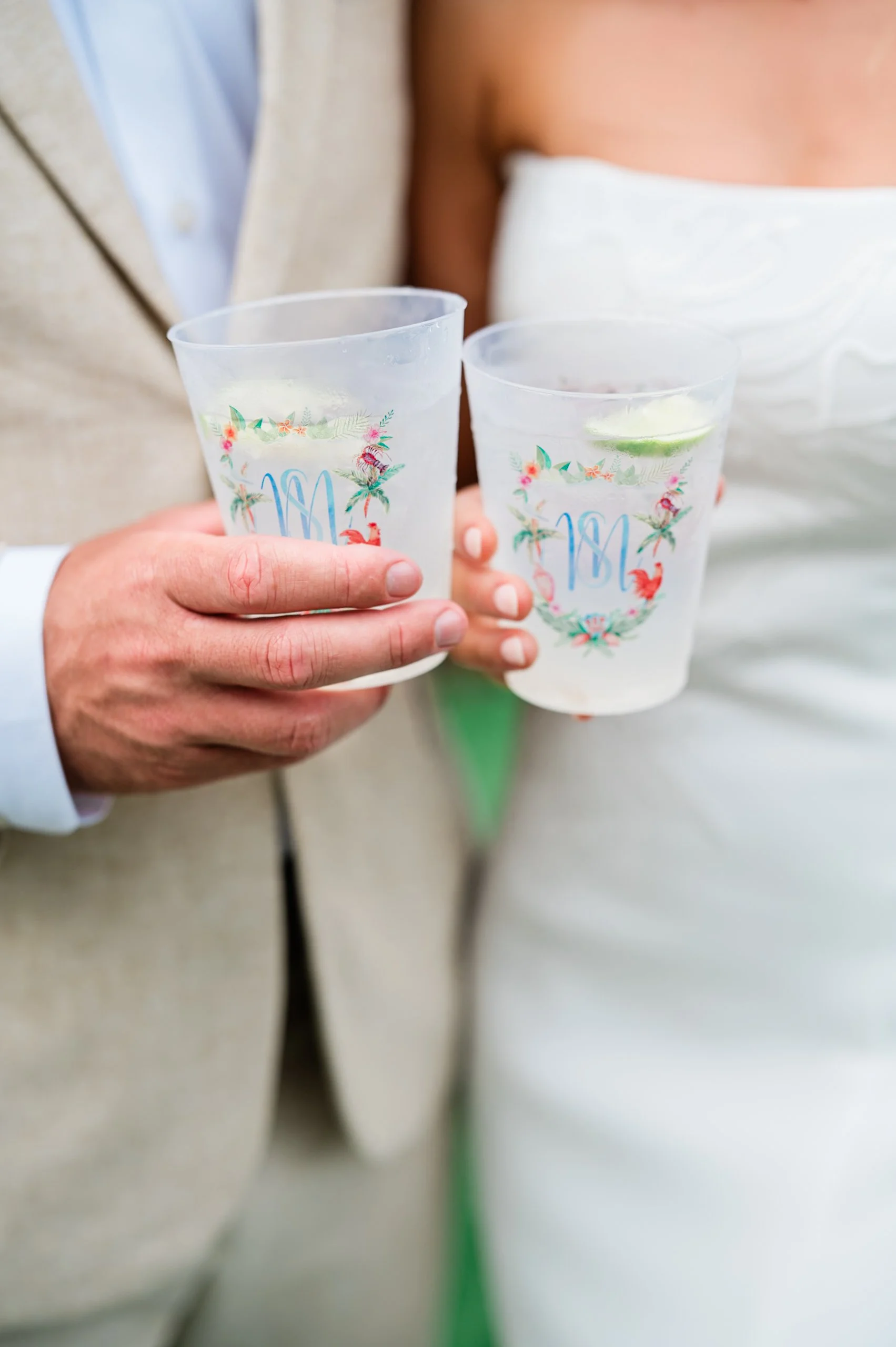 A man and a woman holding clear plastic cups with floral and flamingo designs, containing drinks with lime slices.