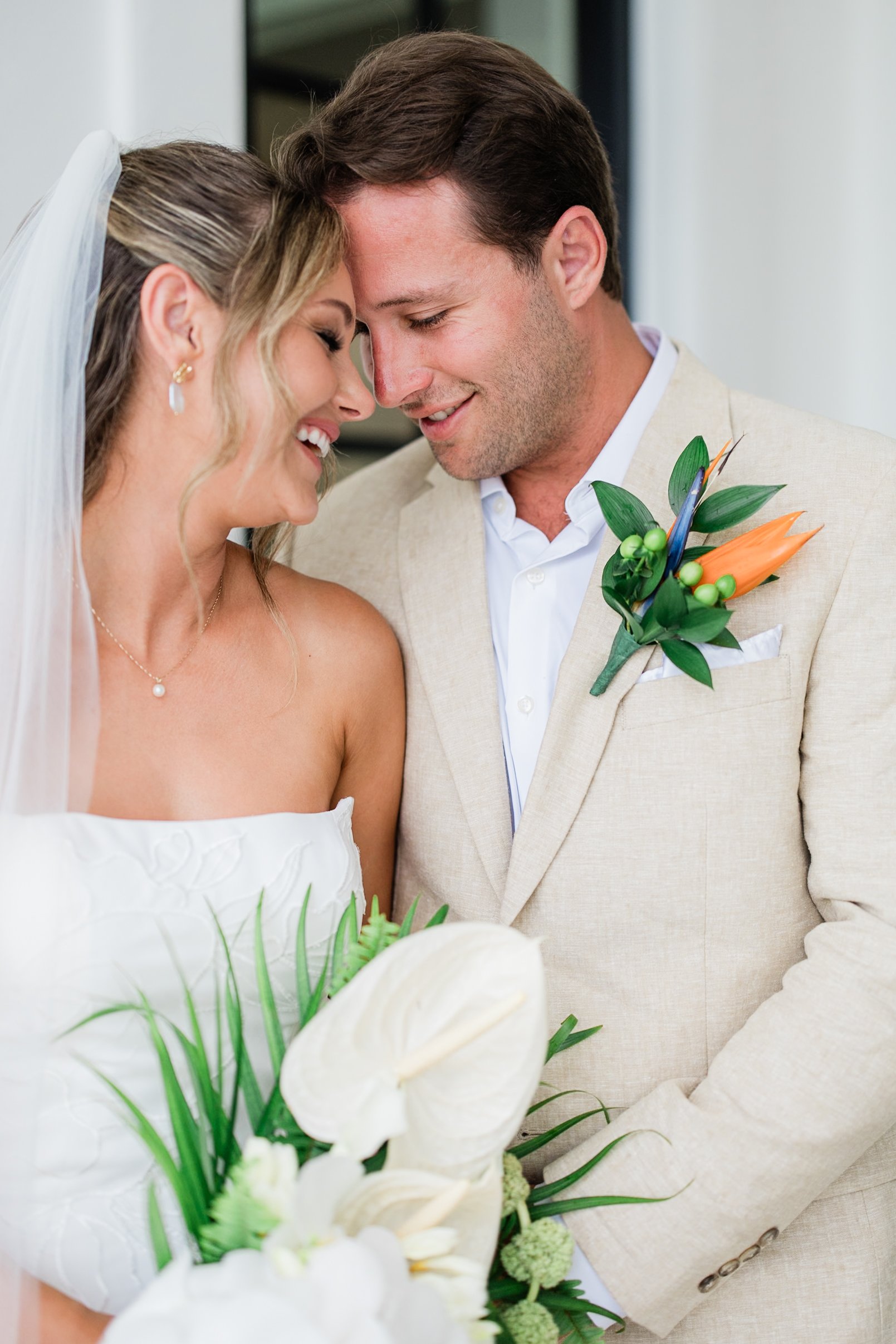 A bride and groom sharing a tender moment with foreheads touching, smiling, during their wedding, with the bride holding a bouquet of white flowers and greenery.