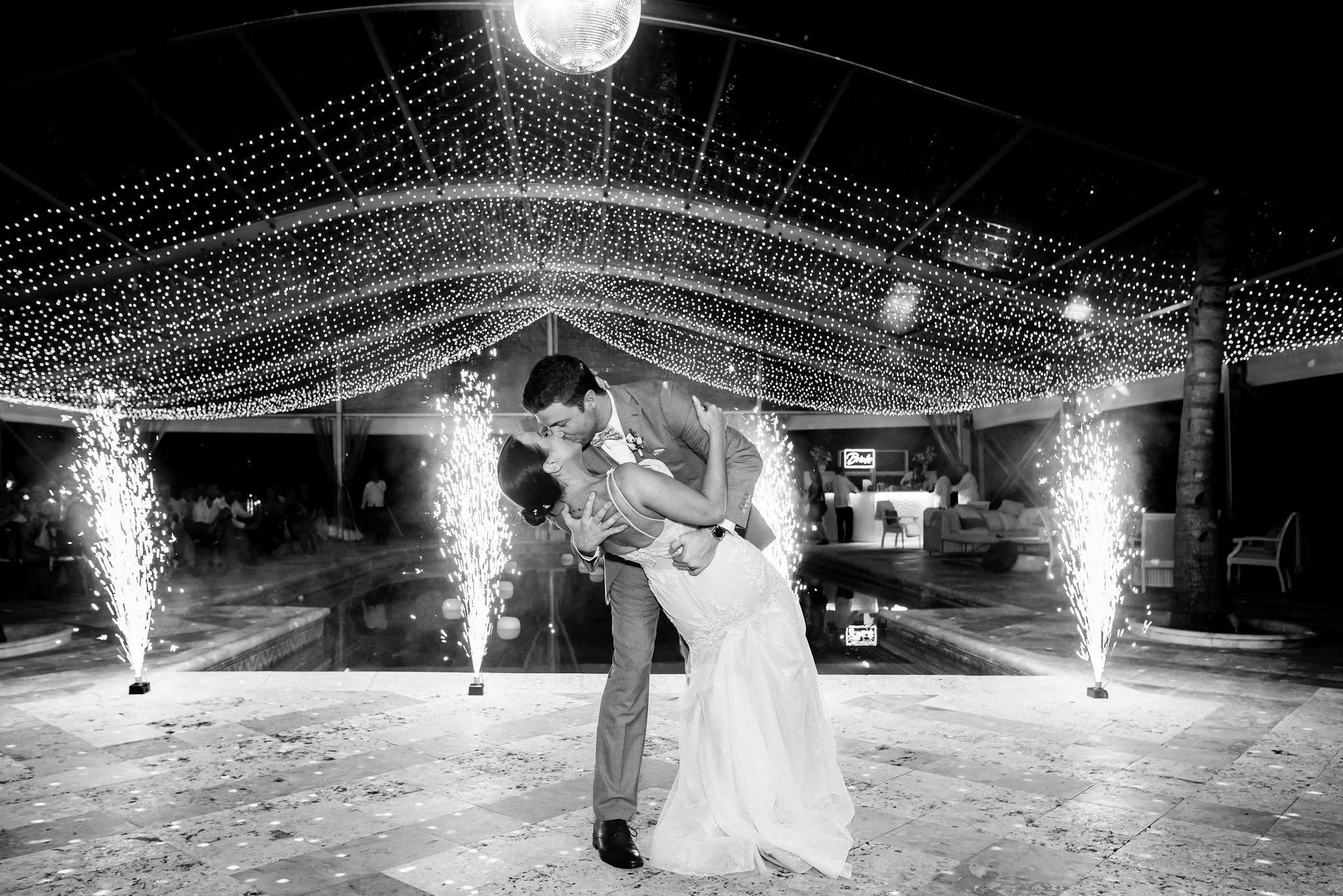 A bride and groom sharing a kiss during their wedding reception under string lights and sparklers.
