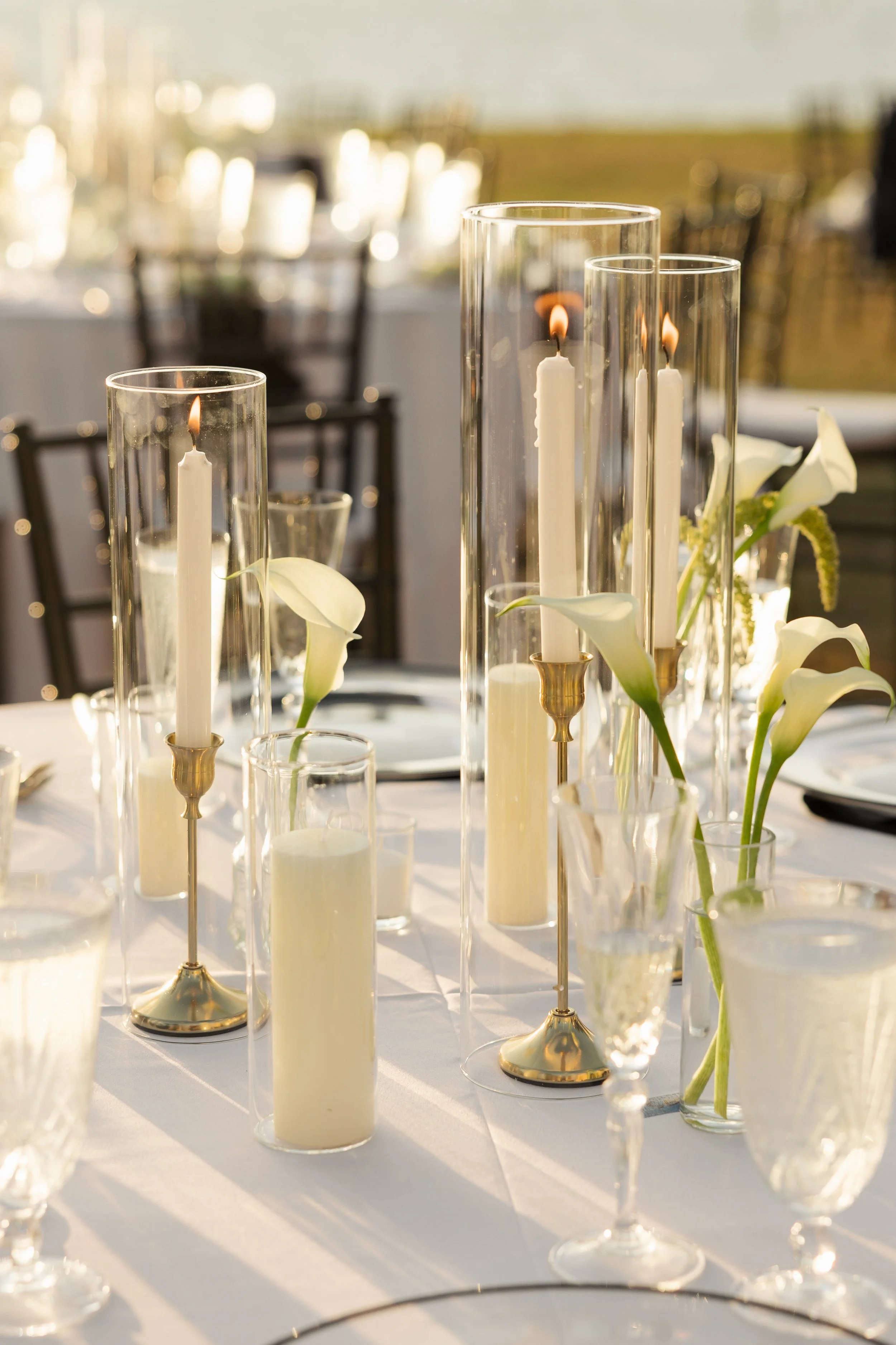 Elegant outdoor dining table decorated with tall glass candle holders, white candles, and white calla lilies, set for a special occasion during sunset.