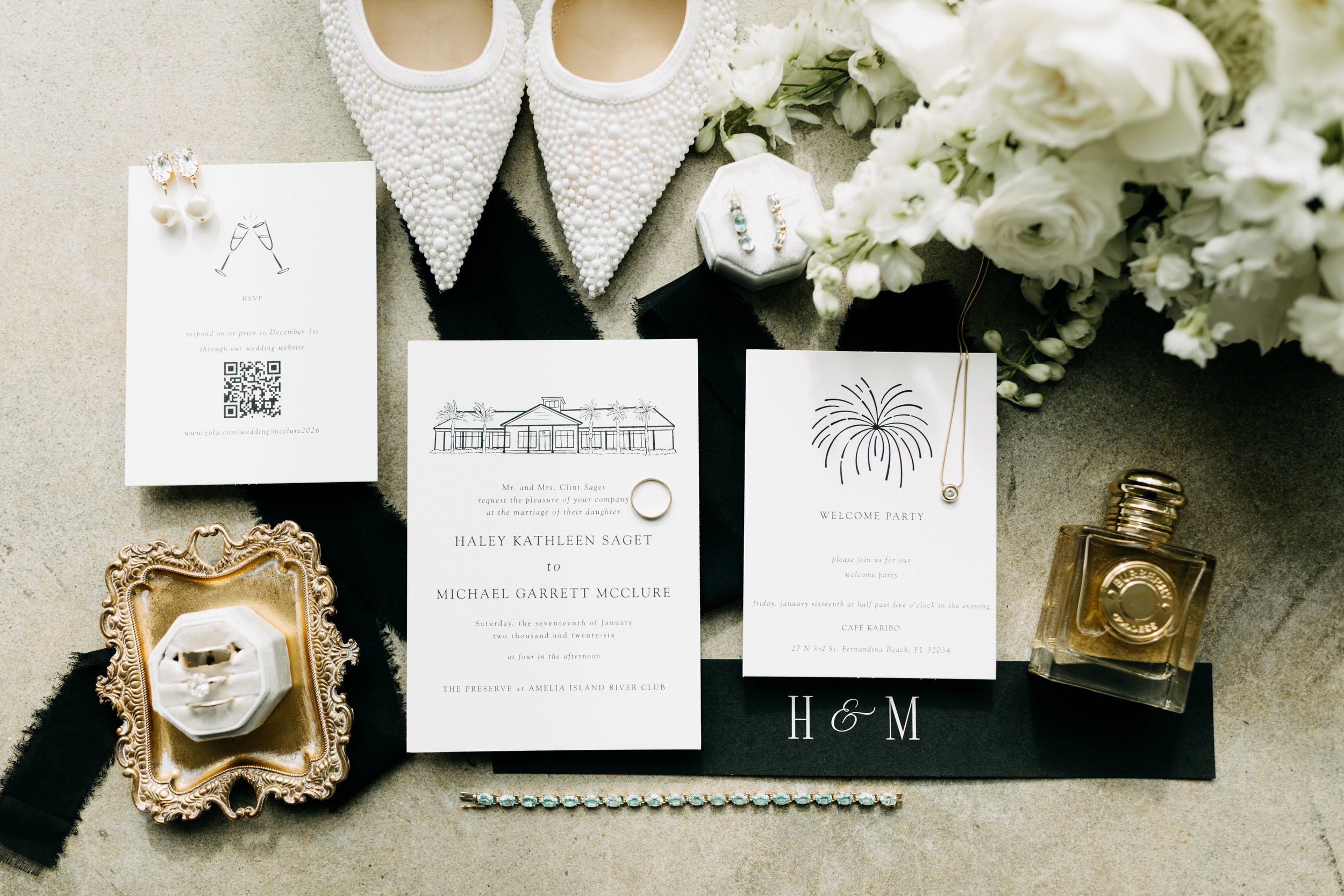 Wedding invitation suite includes a ring, earrings, necklace, perfume, and floral bouquet, with white embellished shoes, a pearl bracelet, and a gold and black tray displayed on a light surface.