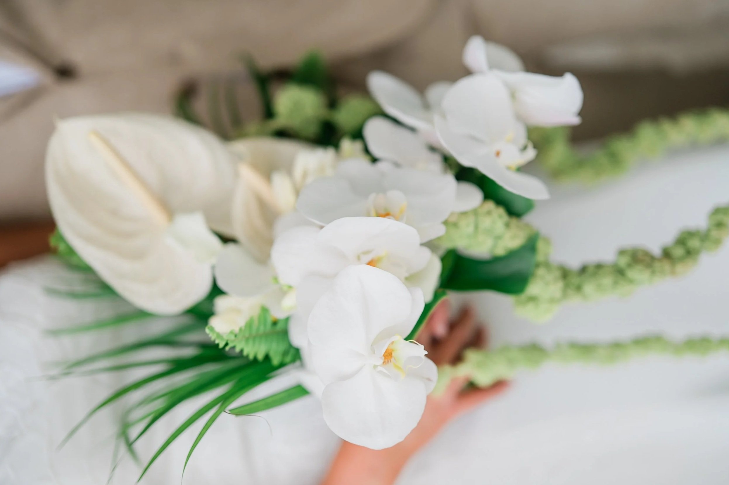 Close-up of a bridal bouquet with white orchids, anthuriums, and greenery, held by a person wearing a white dress.