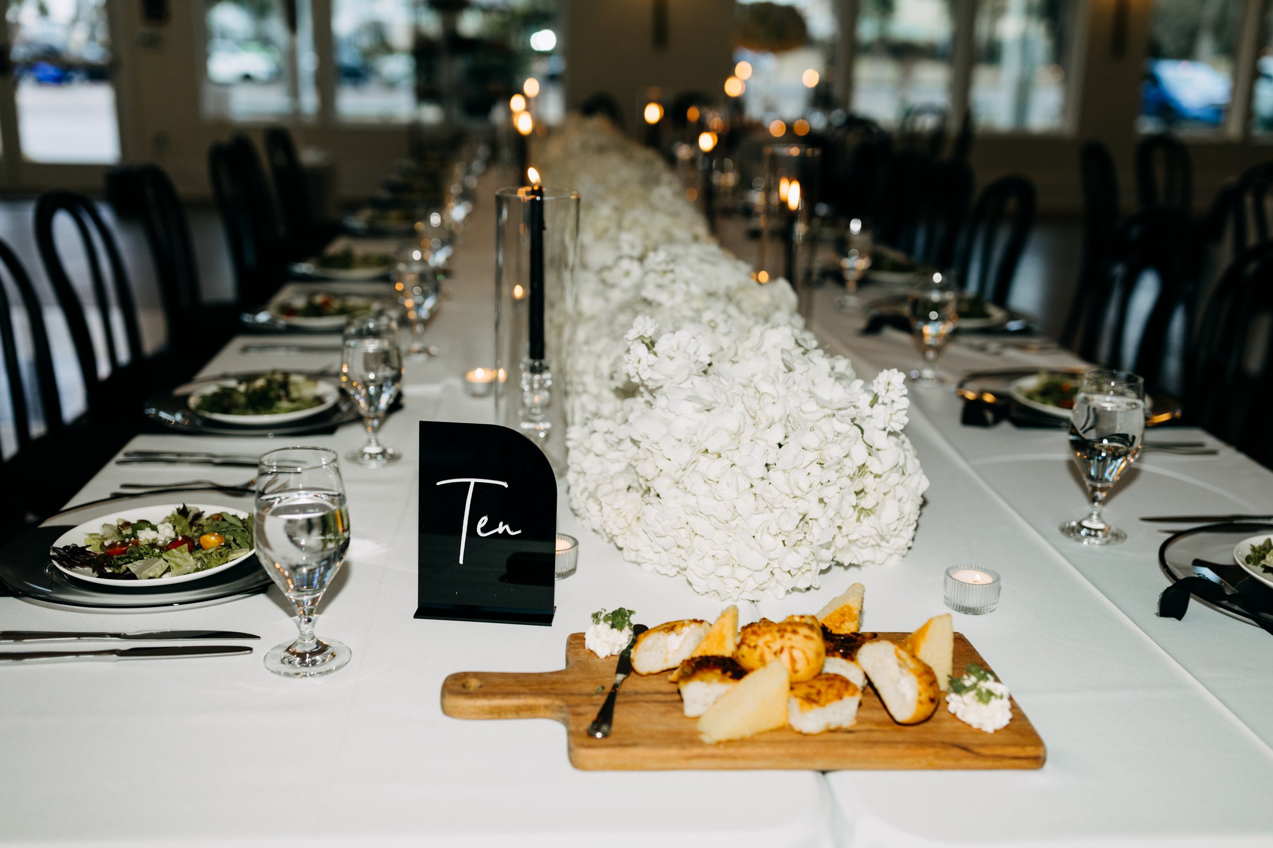 A long dining table set for a celebration with white floral centerpieces, place settings, glasses of water, and candles. There is a black table number card that reads "Ten" and a wooden serving board with slices of cake.