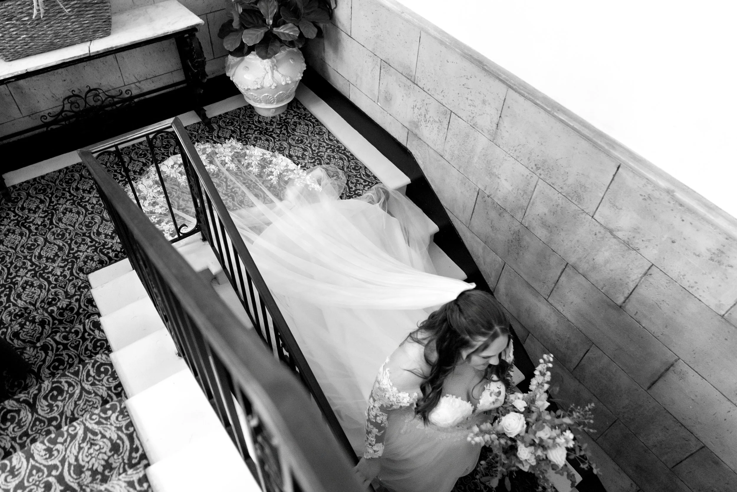 A bride in a wedding dress holding a bouquet of flowers standing on staircase with ornate carpet and stone walls, seen from above.