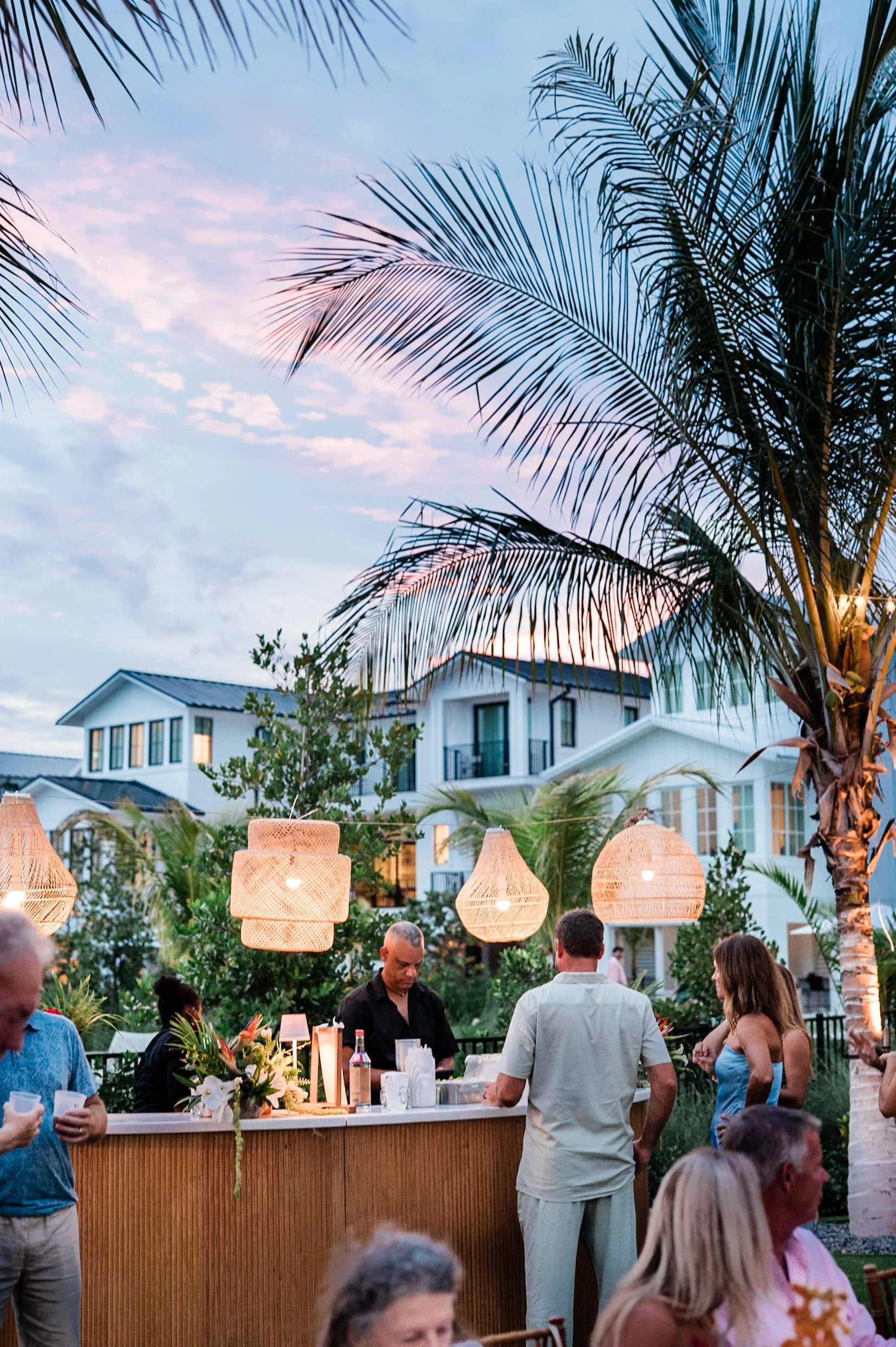 People are at an outdoor gathering with a bar, tropical plants, hanging lanterns, and two-story white houses in the background during sunset.