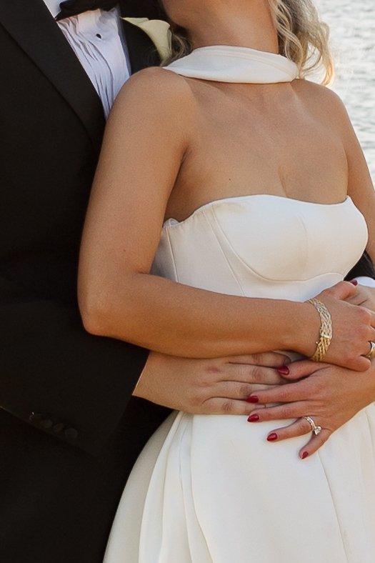 A bride in a strapless white wedding gown and a groom in a black tuxedo holding each other near a body of water.