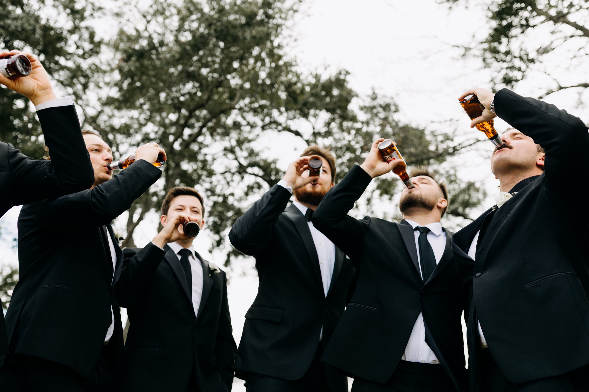 Group of men in tuxedos drinking beer outdoors.
