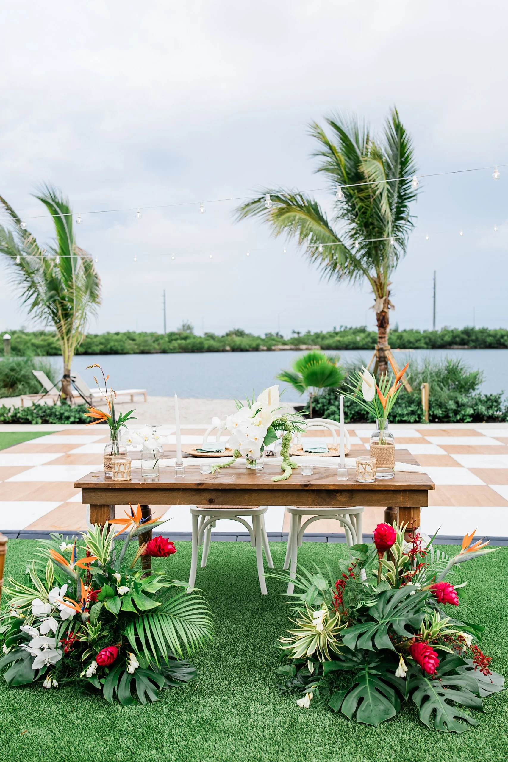 Tropical outdoor event setup featuring a wooden table with floral arrangements, surrounded by lush greenery, on a grassy area near water with palm trees and string lights overhead.