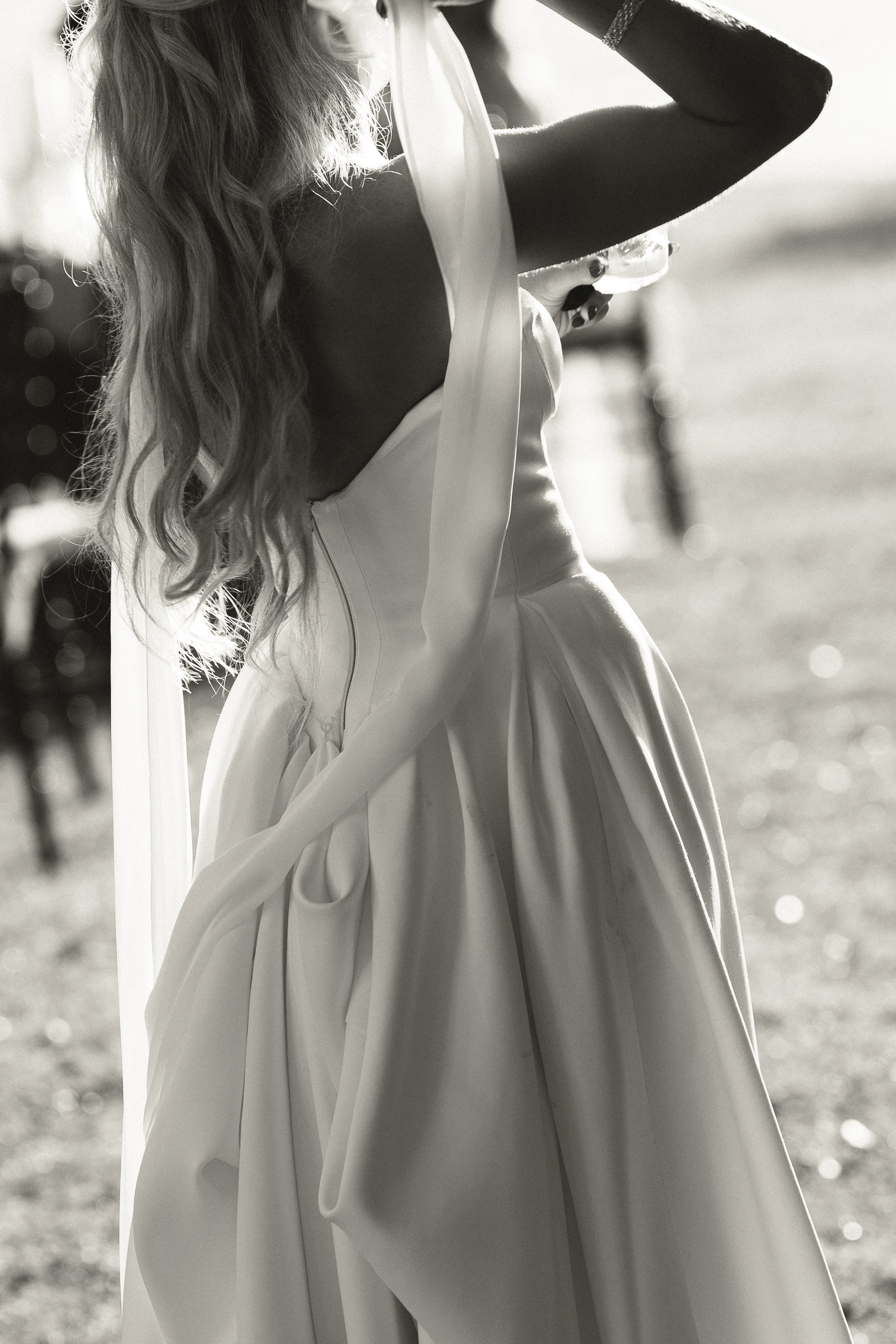 A bride in a wedding dress with long wavy hair stands outdoors, holding her head with one hand, with sunlight illuminating her and a blurred background of a person and trees.