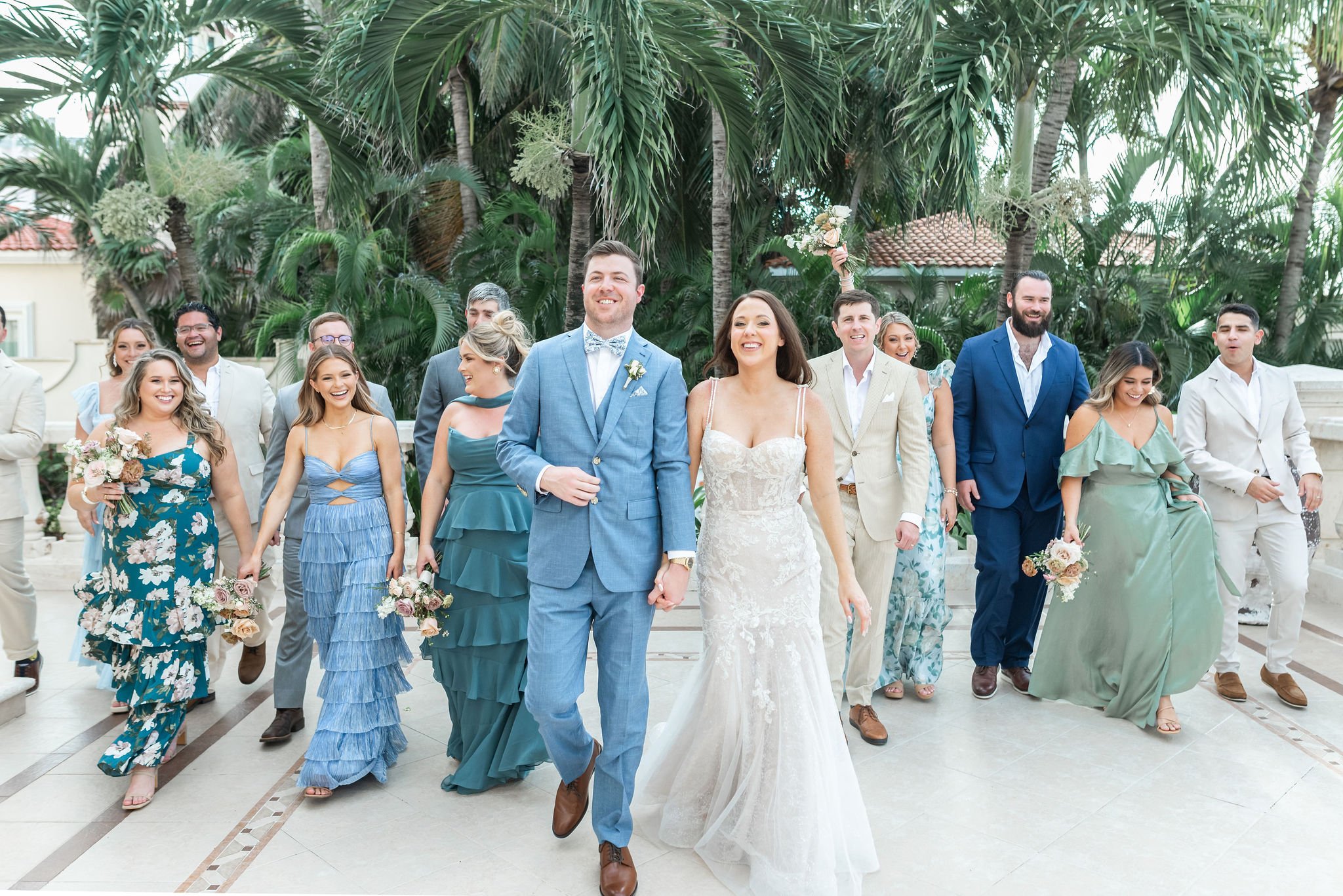 A group of wedding guests walking outdoors, with the bride and groom in front, under palm trees.