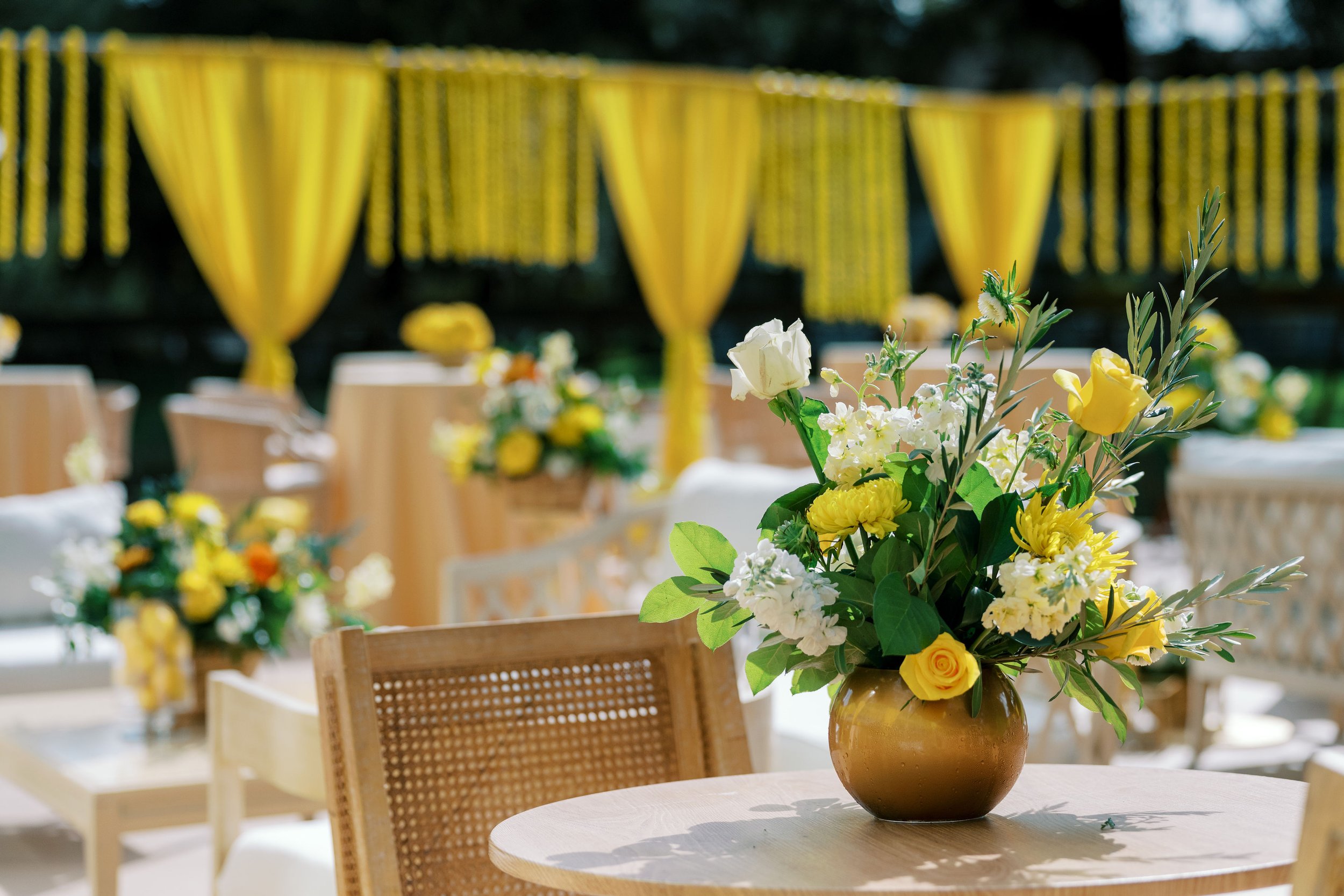 A flower arrangement with yellow and white flowers on a wooden table at an outdoor event.