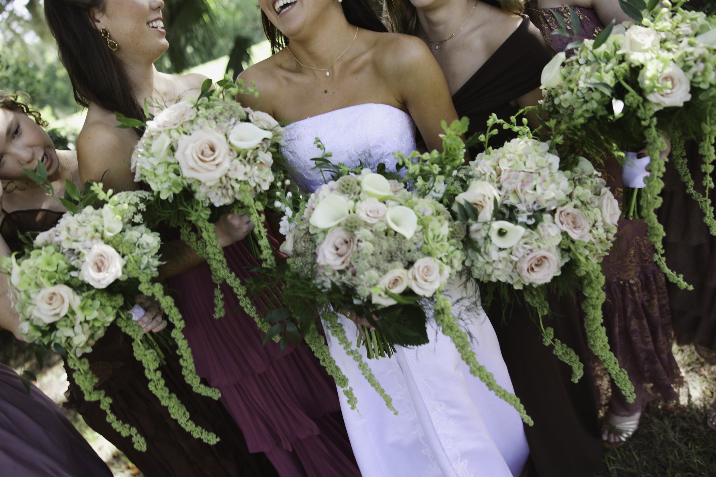 Group of women dressed in bridal and bridesmaid gowns holding bouquets of pink and white flowers at an outdoor wedding.