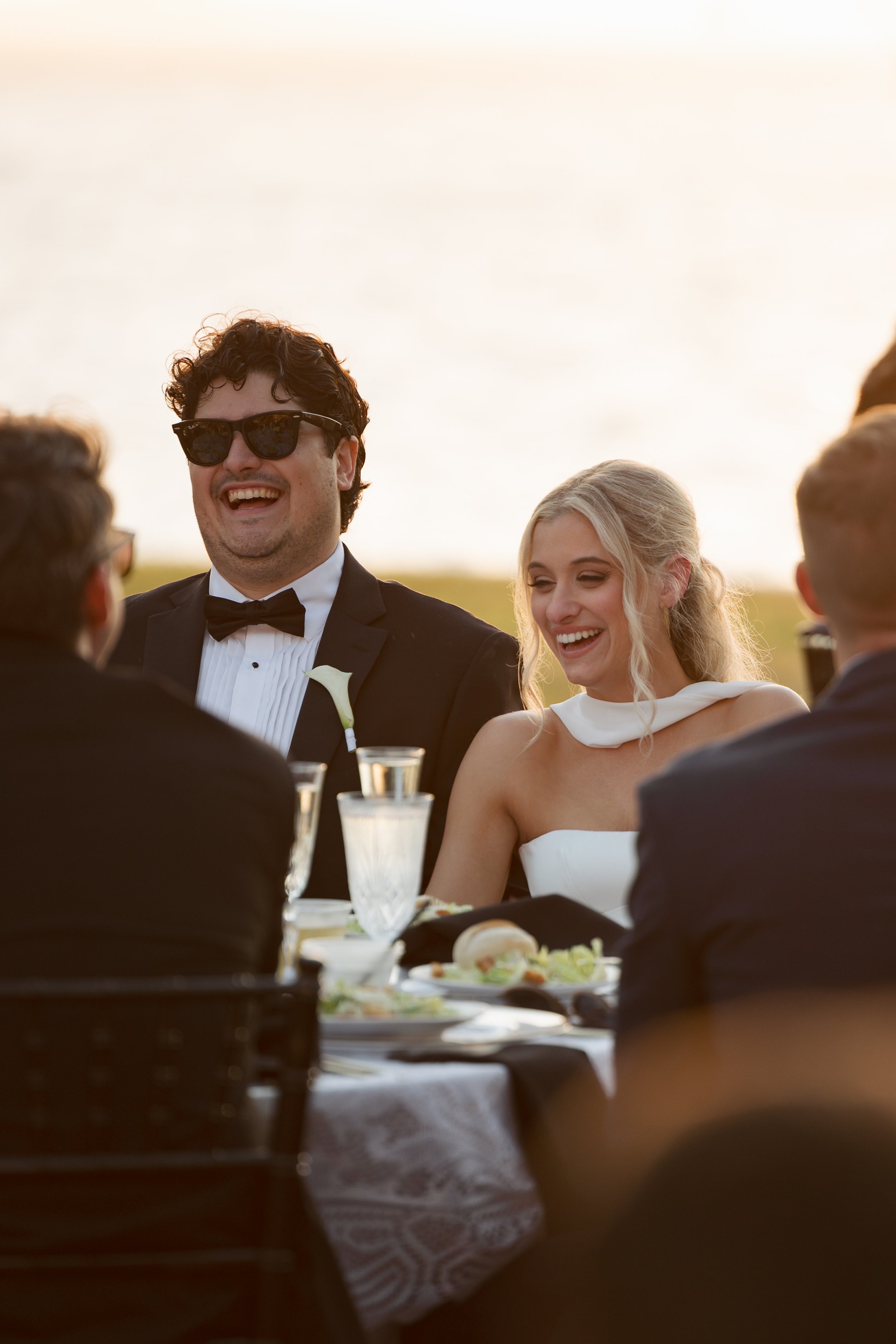 A group of people, including a man in a tuxedo and sunglasses and a woman in a white dress, enjoying a meal outdoors during sunset at a wedding reception.