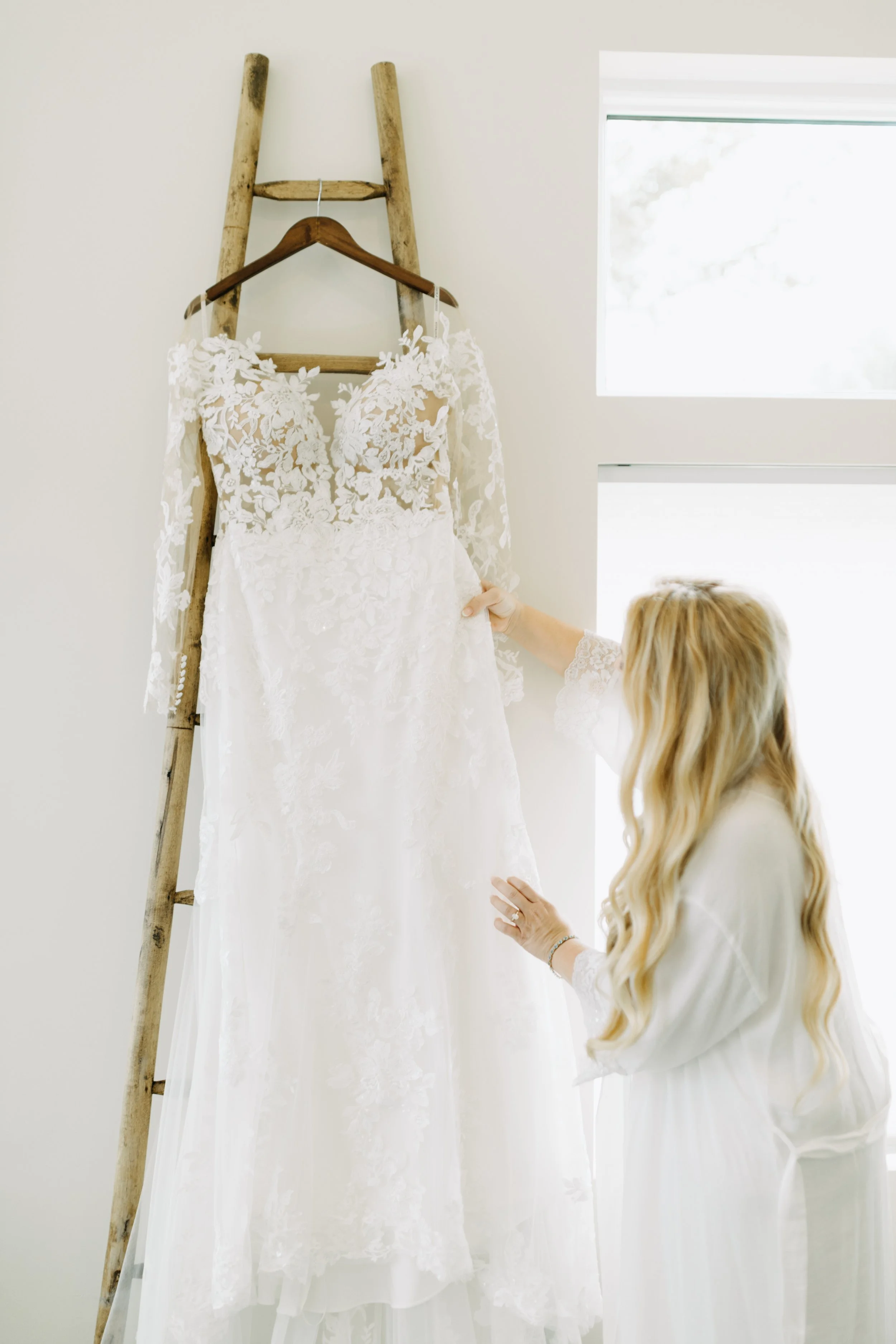Bridesmaid in white dress holding a wedding gown with lace details, hanging on a wooden ladder against a white wall near a window.
