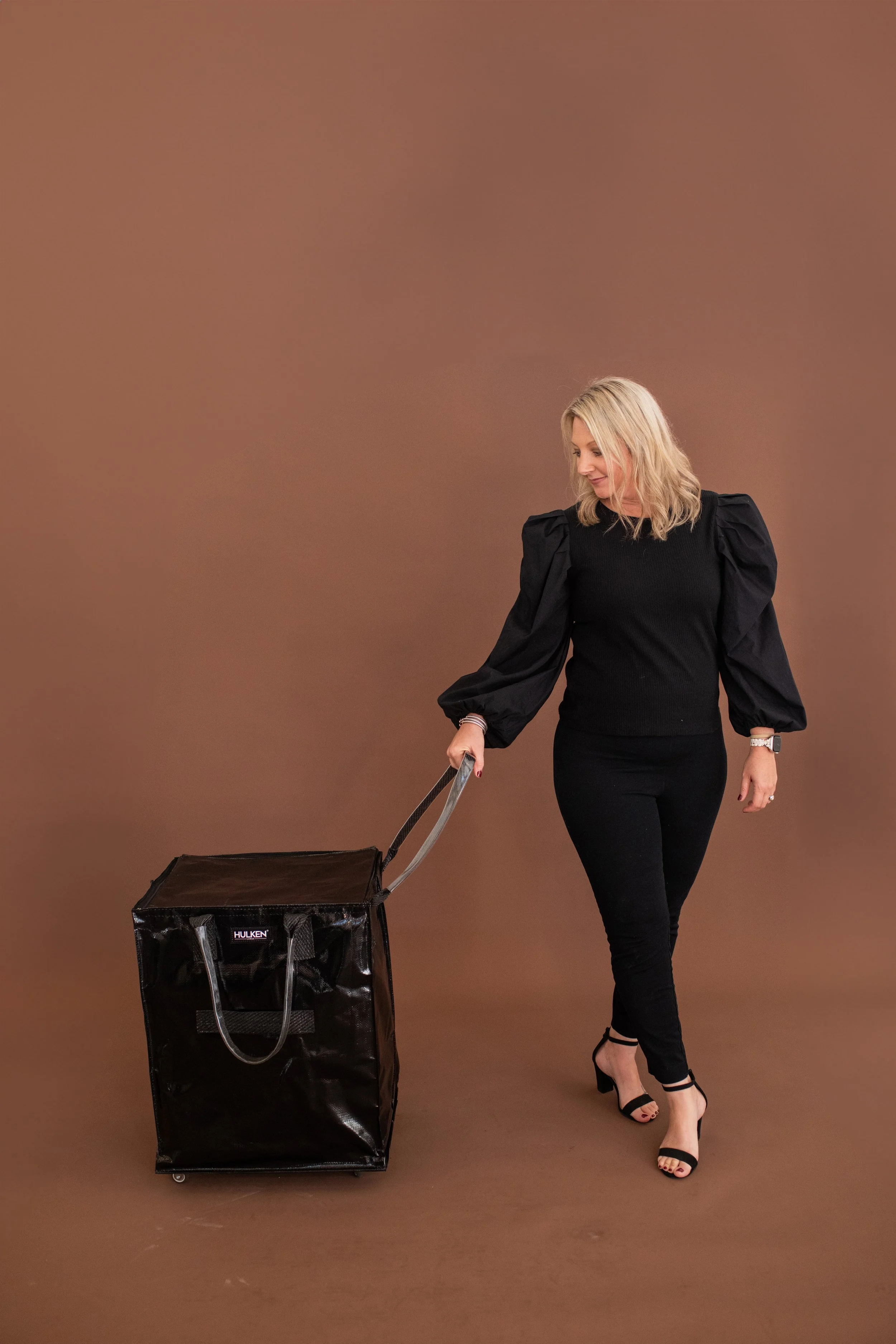 Woman with blonde hair in black outfit pulling a black insulated delivery bag on wheels.