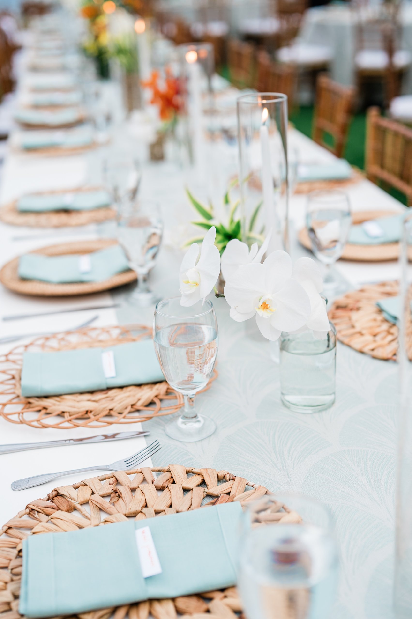 Elegant outdoor dining table decorated with white orchids, tall glass vases, and candles, set with woven placemats, light blue napkins, water glasses, and silverware.