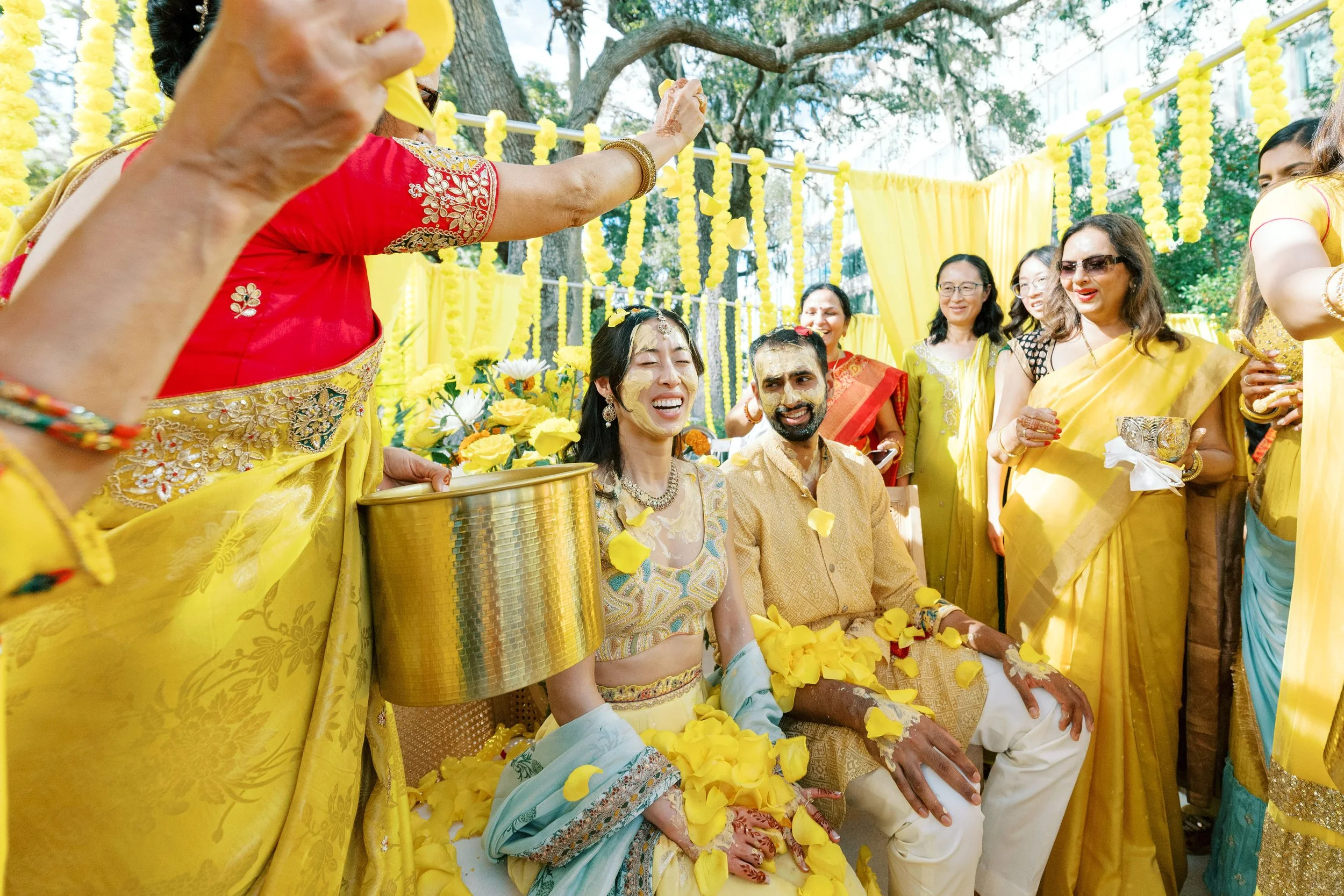 Indian wedding ceremony outdoors with yellow decorations and flower petals, featuring a bride and groom sitting together surrounded by women in traditional yellow sarees, one of whom is pouring flower petals over them.