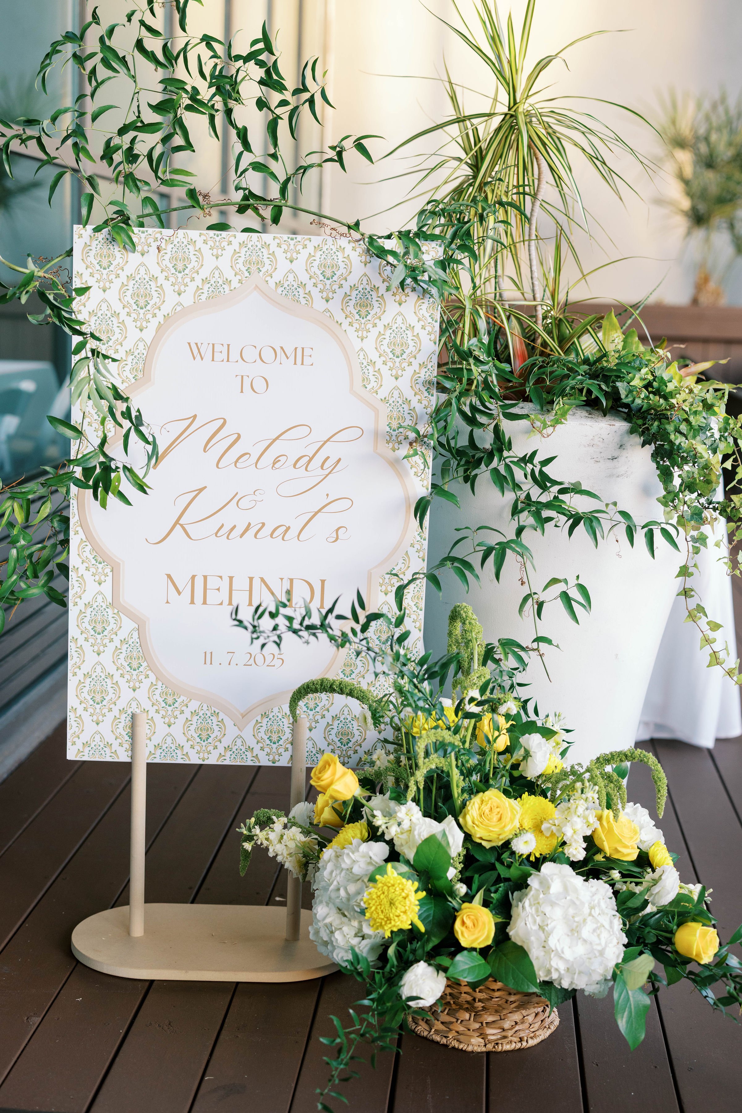 A decorative welcome sign with gold and pastel patterns surrounded by lush green plants and a basket of yellow and white flowers, placed on a wooden surface, likely for a wedding or celebration.