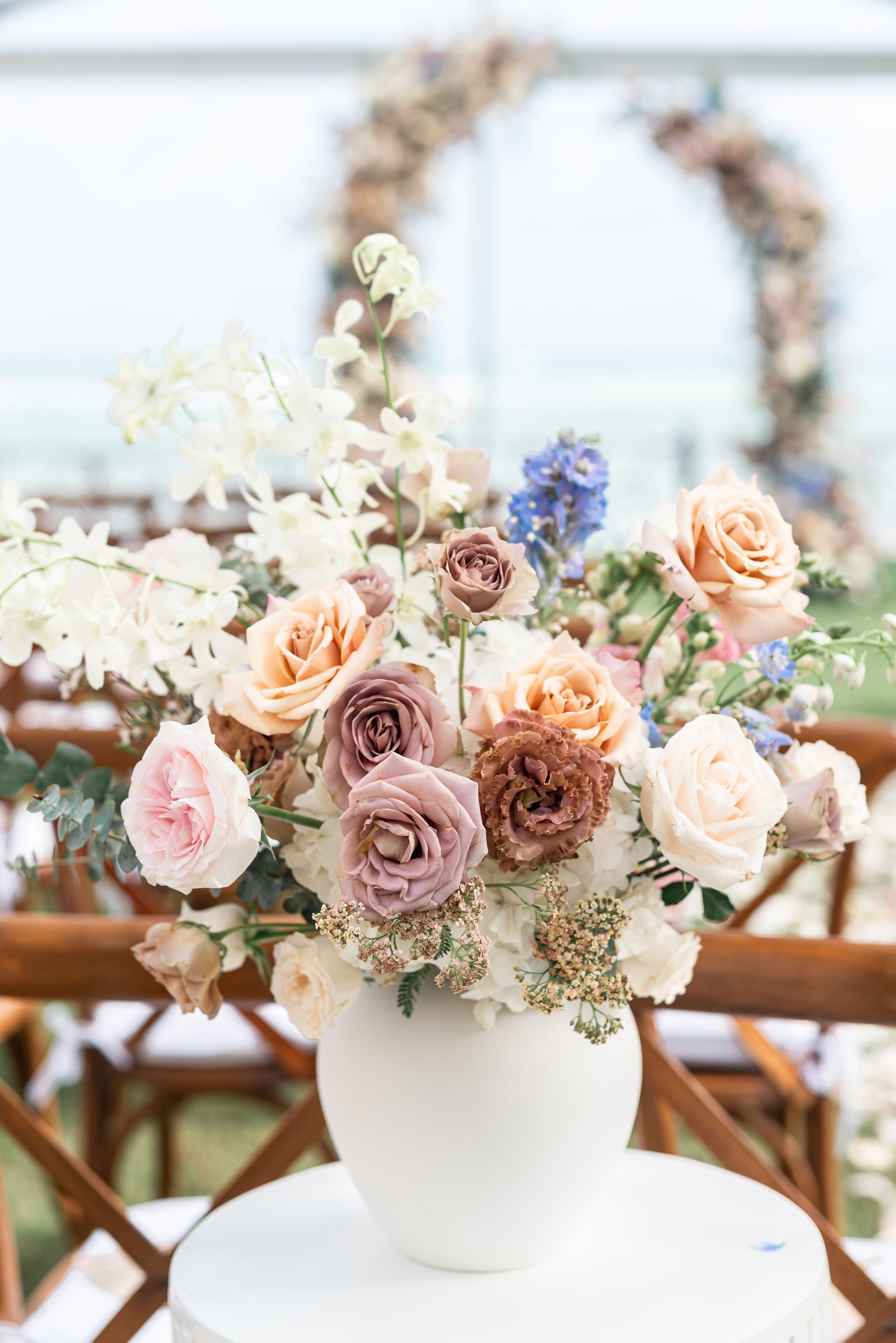 A large floral arrangement with roses, orchids, and other flowers in a white vase on a round white table.