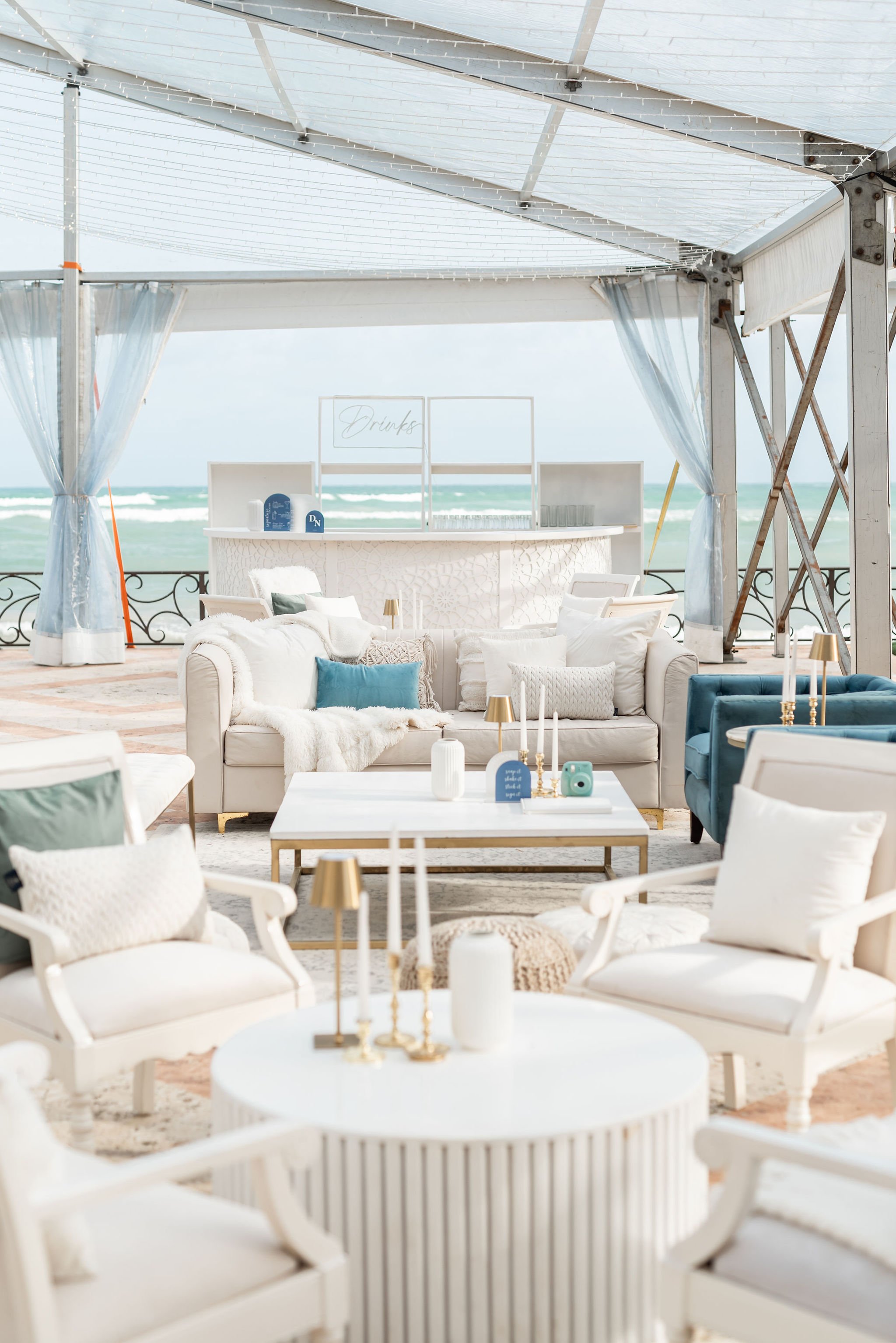 Beachside outdoor lounge with white and blue furniture, candles, and a bar area with a "Drinks" sign, overlooking the ocean under a canopy with string lights.