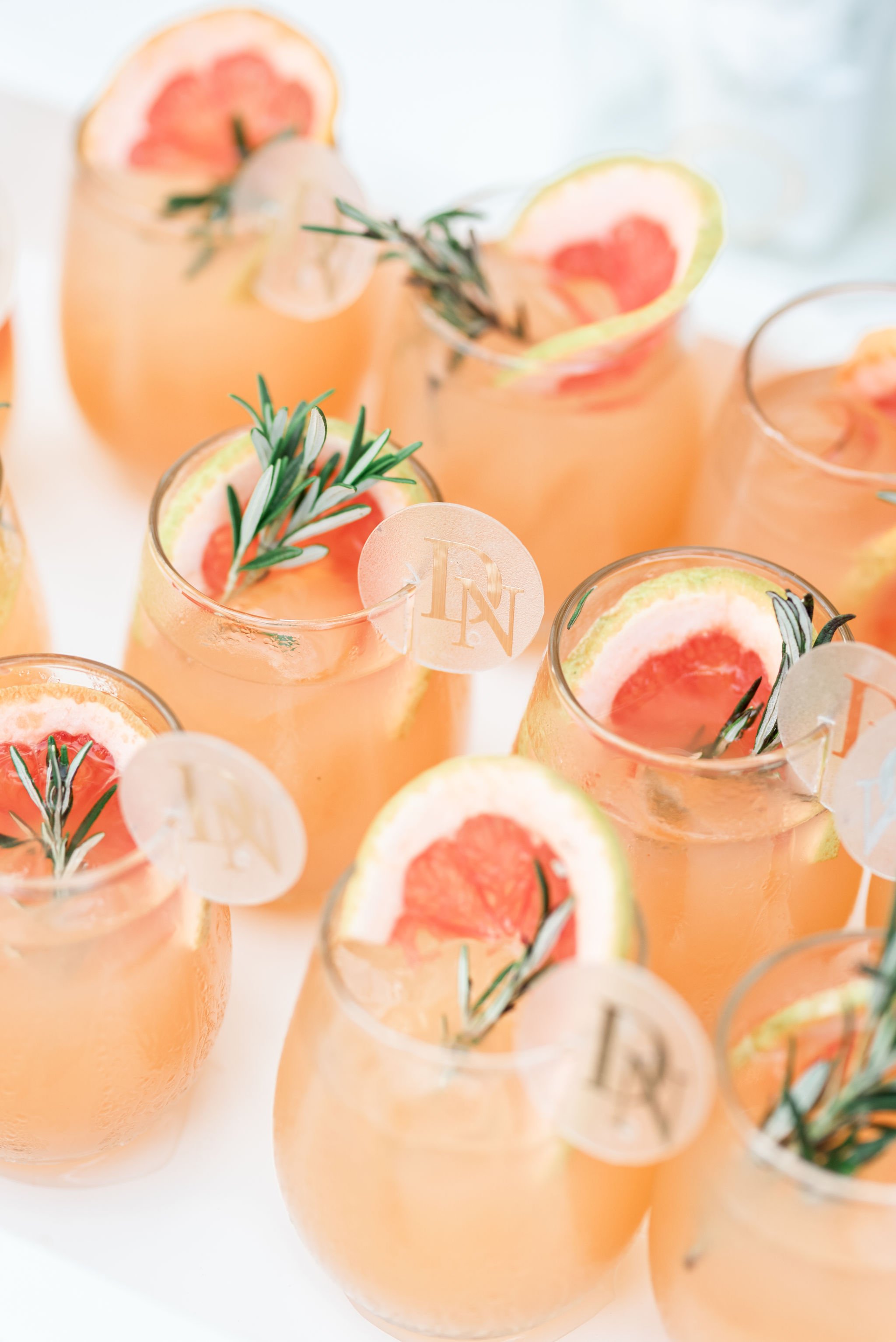 Several glasses of pink grapefruit cocktails garnished with grapefruit slices and sprigs of rosemary.