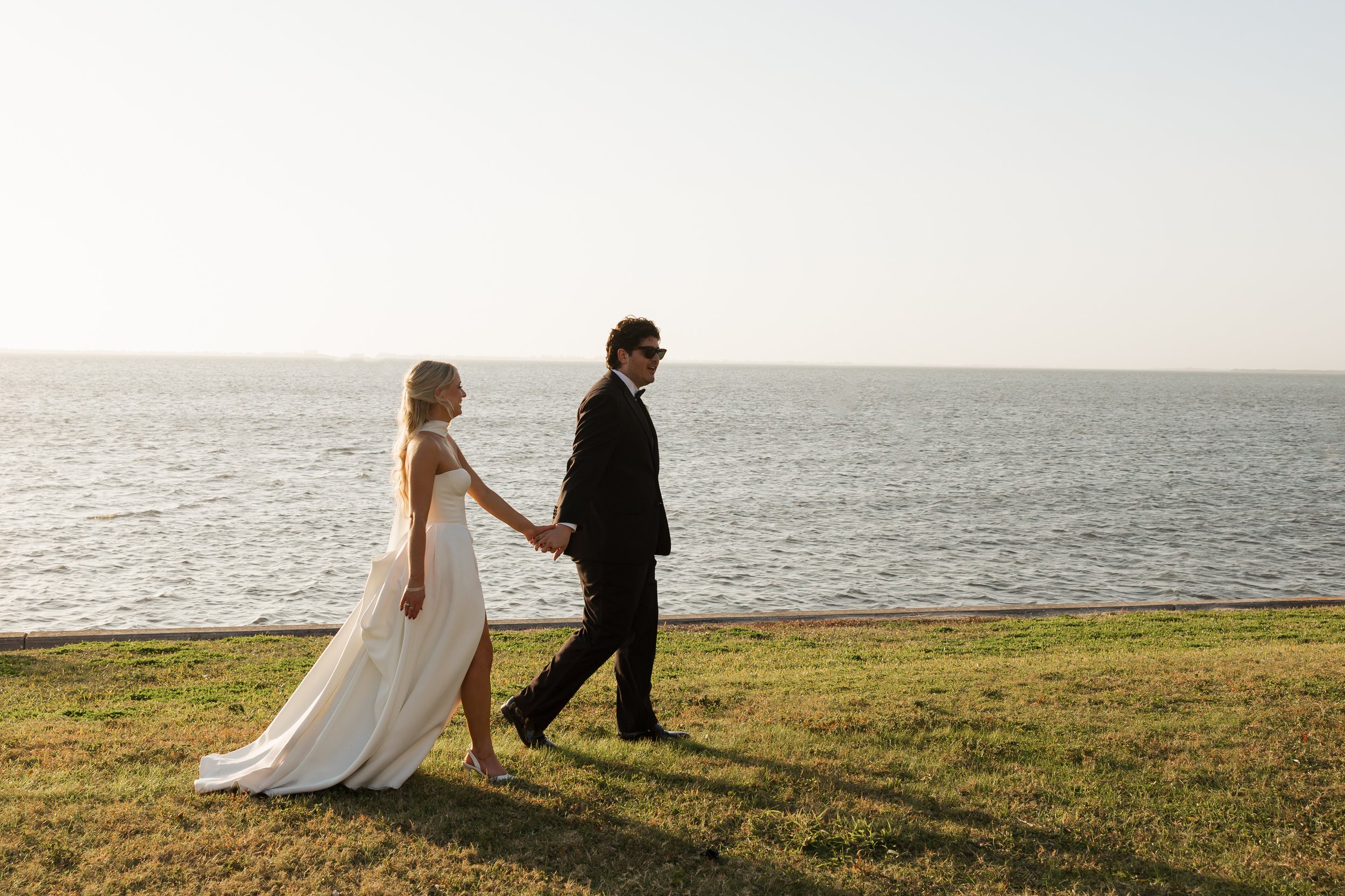 A bride and groom holding hands and walking along a grassy area near a body of water at sunset.