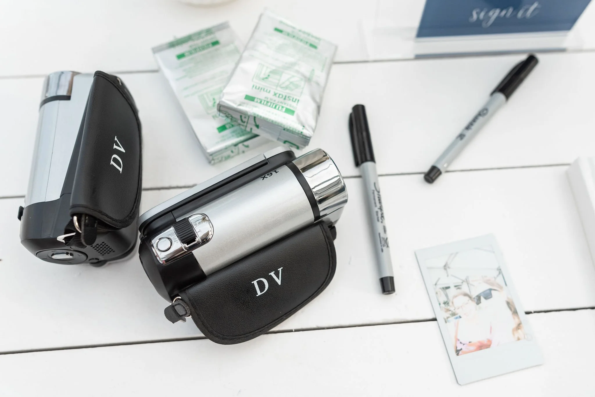 Two portable breathalyzer devices labeled 'DV' on a white surface, with two pens, a packet of tissues, sunglasses, a small photo, and a sign in the background.