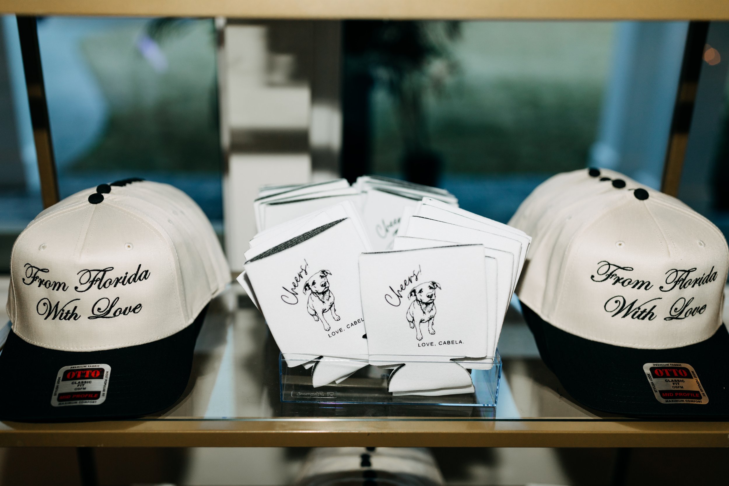 Two white and black baseball caps with embroidery that says 'From Florida With Love.' In front of the caps is a clear box containing Fido-themed stationery or cards with a cartoon dog and the words 'Cheers!' and 'Love, Cabela.'